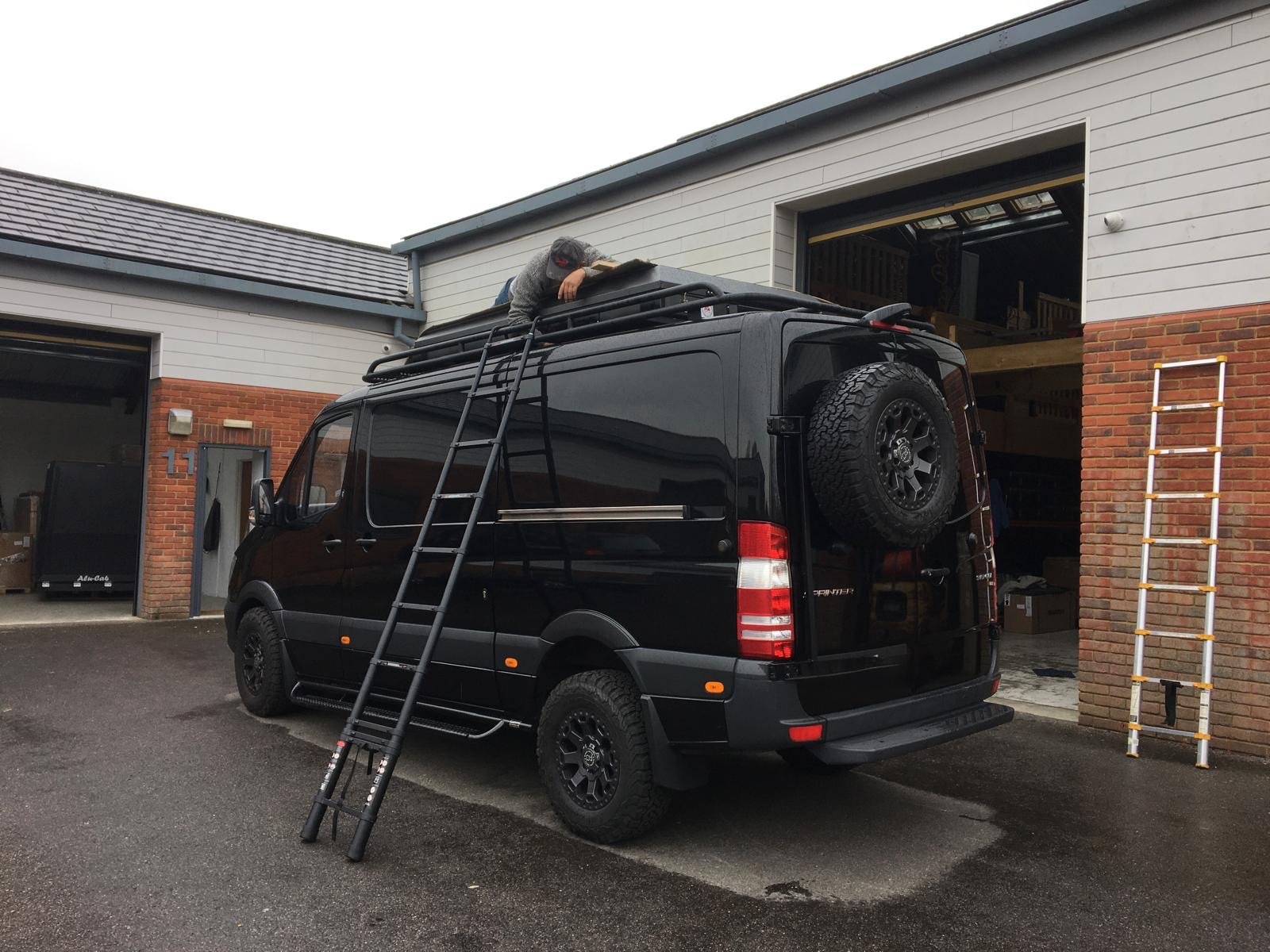 A person on a ladder leaning on the roof of a black van, parked outside a garage, with another ladder standing on the right side of the garage.