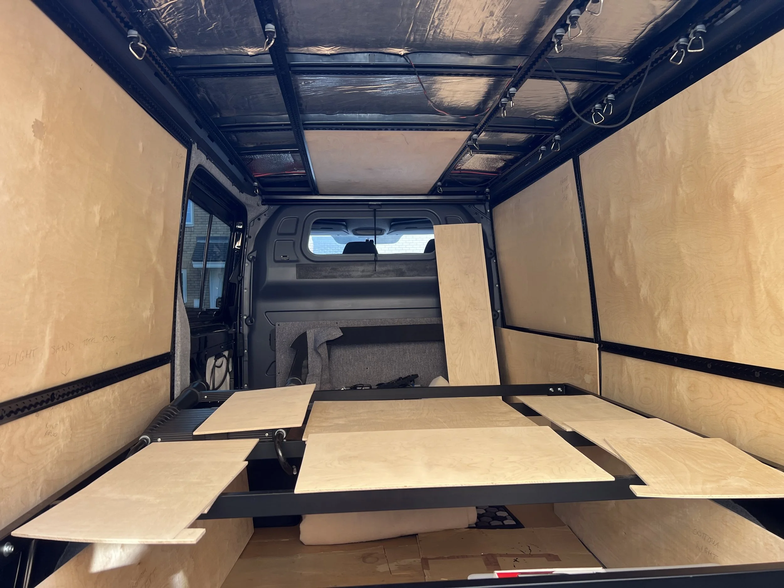Interior of a van being converted with plywood panels on the walls and ceiling, wooden shelves, and a partially installed wooden platform or bed frame.