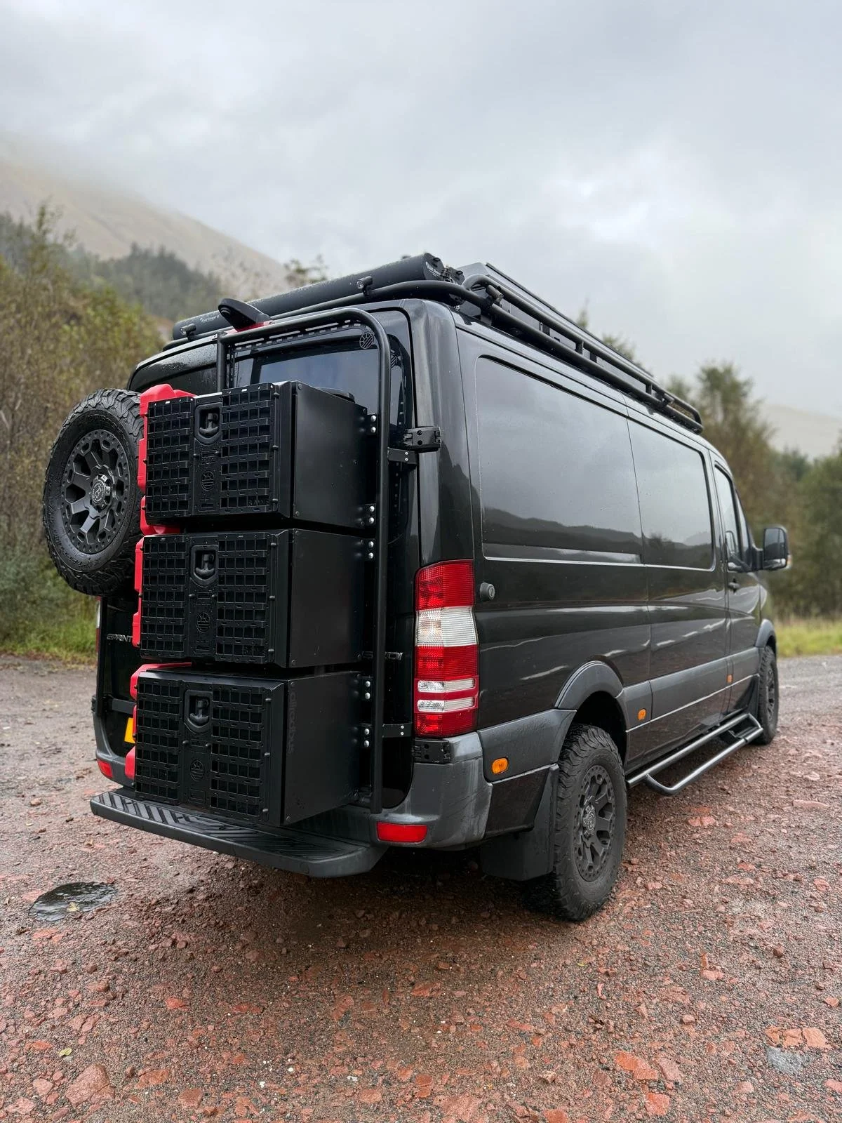 Black van parked outdoors with equipment and spare tire mounted on the rear, set against a cloudy sky and mountainous background.