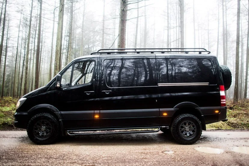 A black van parked on a dirt road in a foggy forest with tall trees