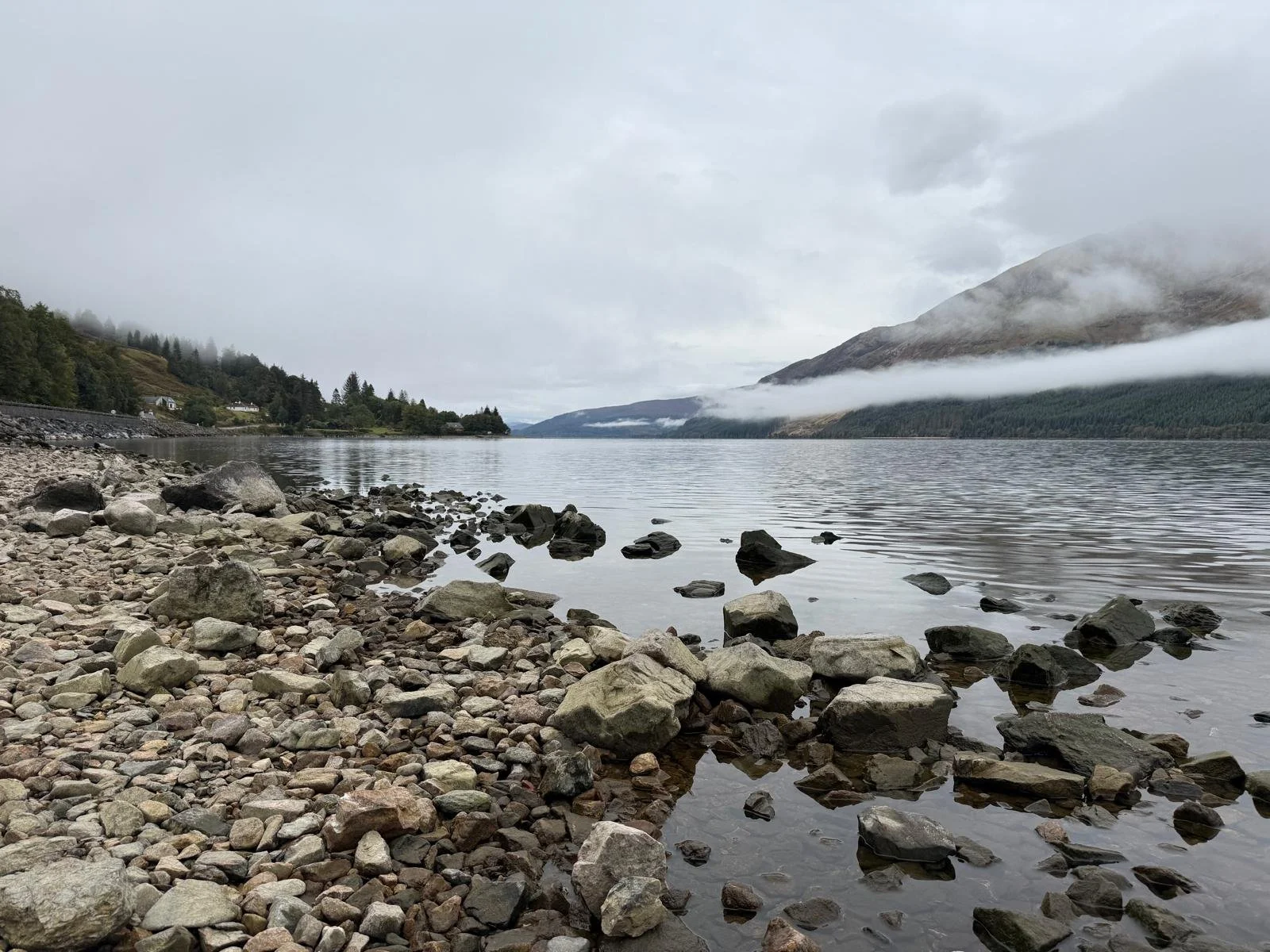 A rocky lakeshore with calm water, surrounded by green hills and mountains partially covered by fog or low clouds.