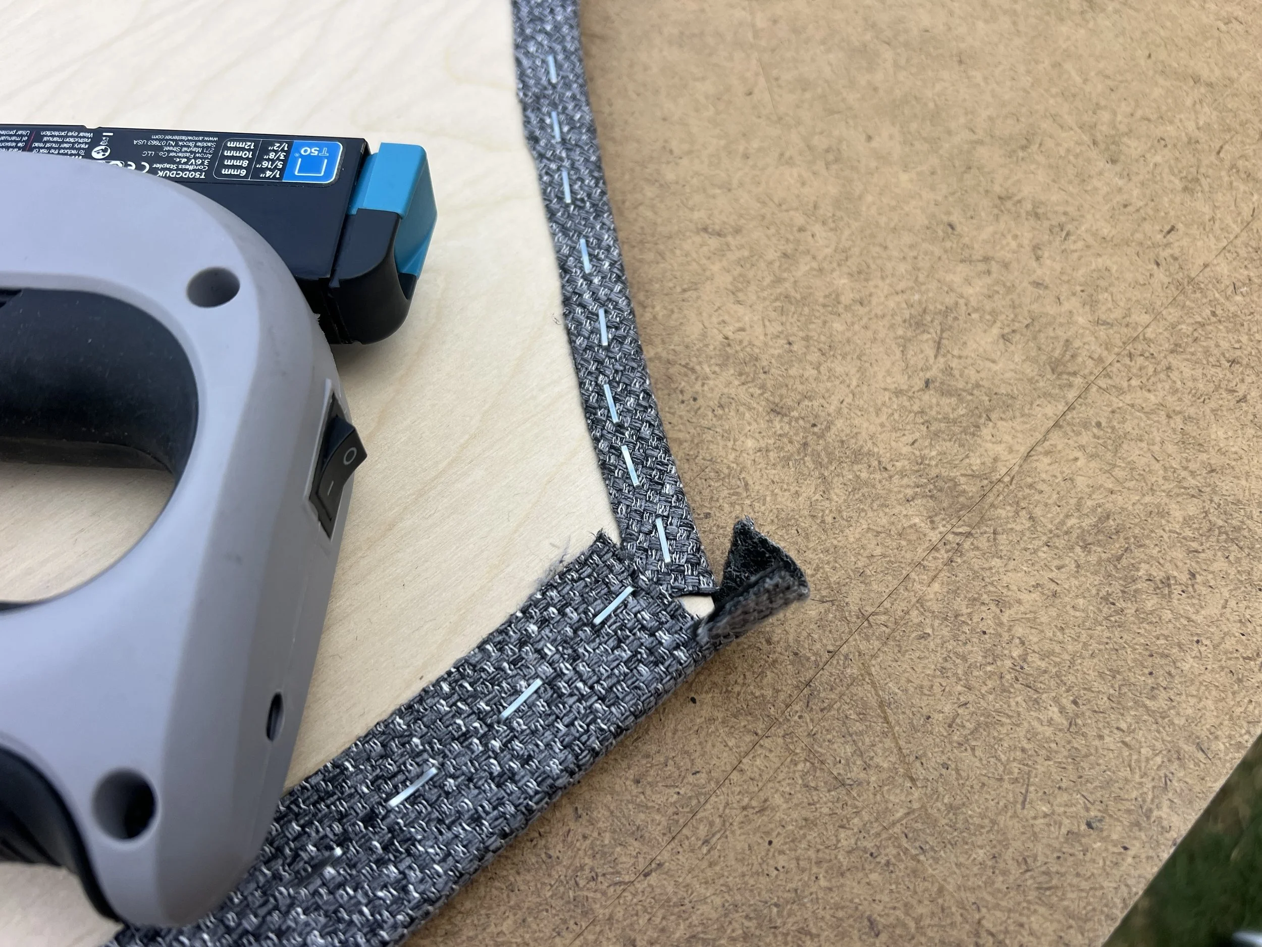 Close-up of a corner of a work surface, showing a cordless drill, a piece of fabric, and a stapler.