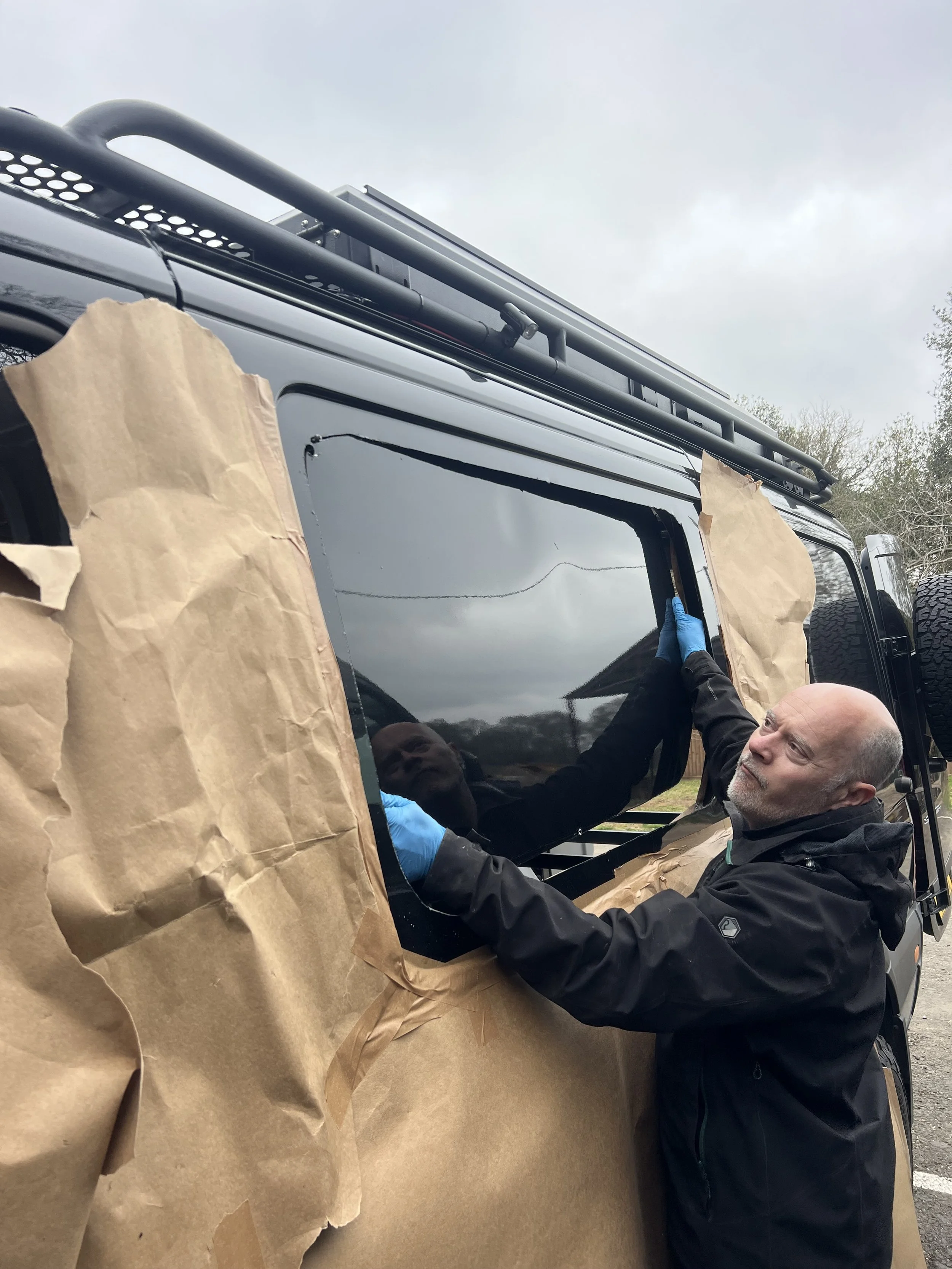 A man in a black jacket and blue gloves is working on the window of a black vehicle, with brown paper covering the surrounding area, on a cloudy day.
