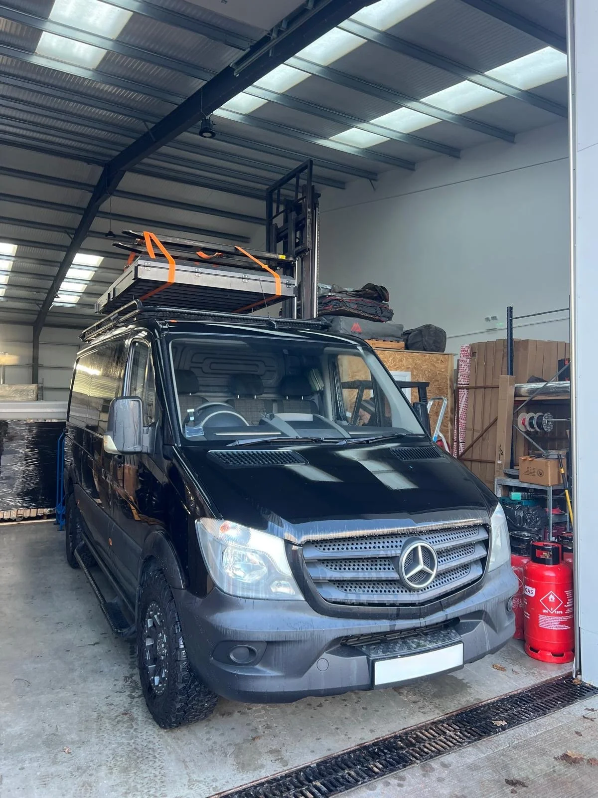 Black Mercedes-Benz van parked inside a warehouse or garage, with equipment and supplies stored around it.