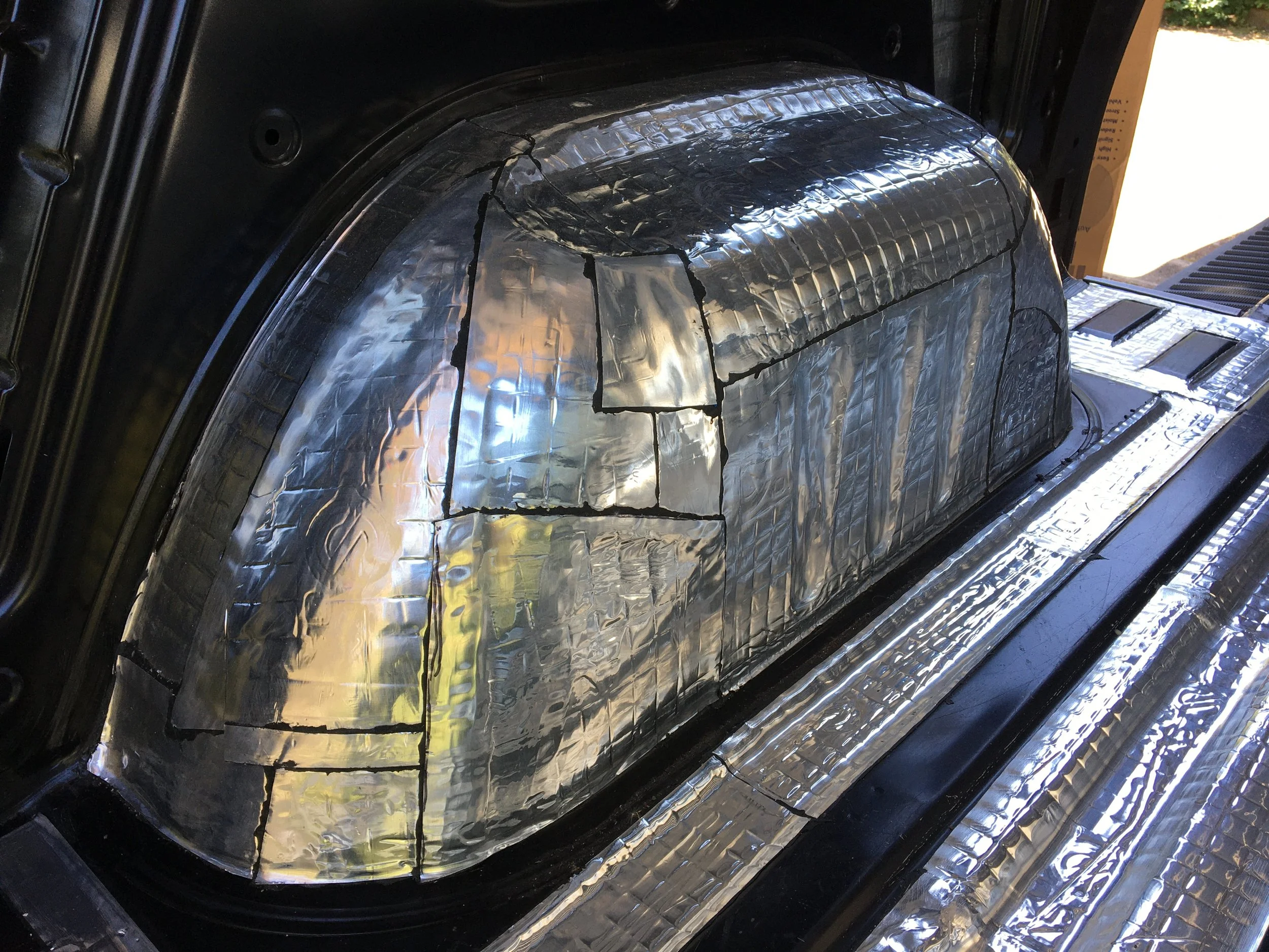A section of a vehicle interior with a black, shiny surface covered with silver reflective foil insulation, with visible seams and folds.