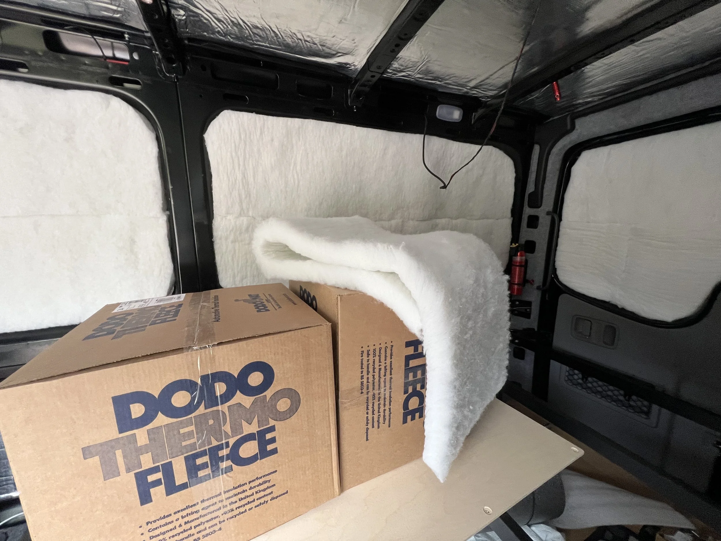 Car interior with partially installed white thermal insulation and a cardboard box labeled "DODO Thermo Fleece" on a surface inside