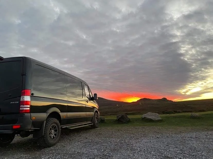 A black van parked on a gravel area with a scenic landscape of hills and rocks under a cloudy sky at sunset.