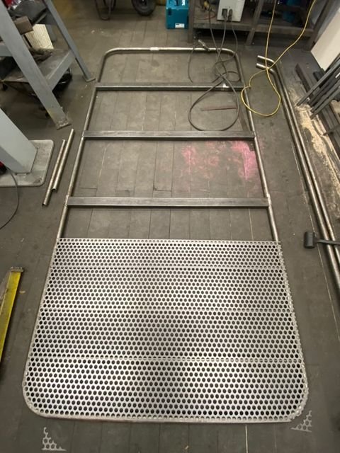 Metal grid platform with a rounded front and a rectangular frame, lying on a workshop floor with tools and equipment around.