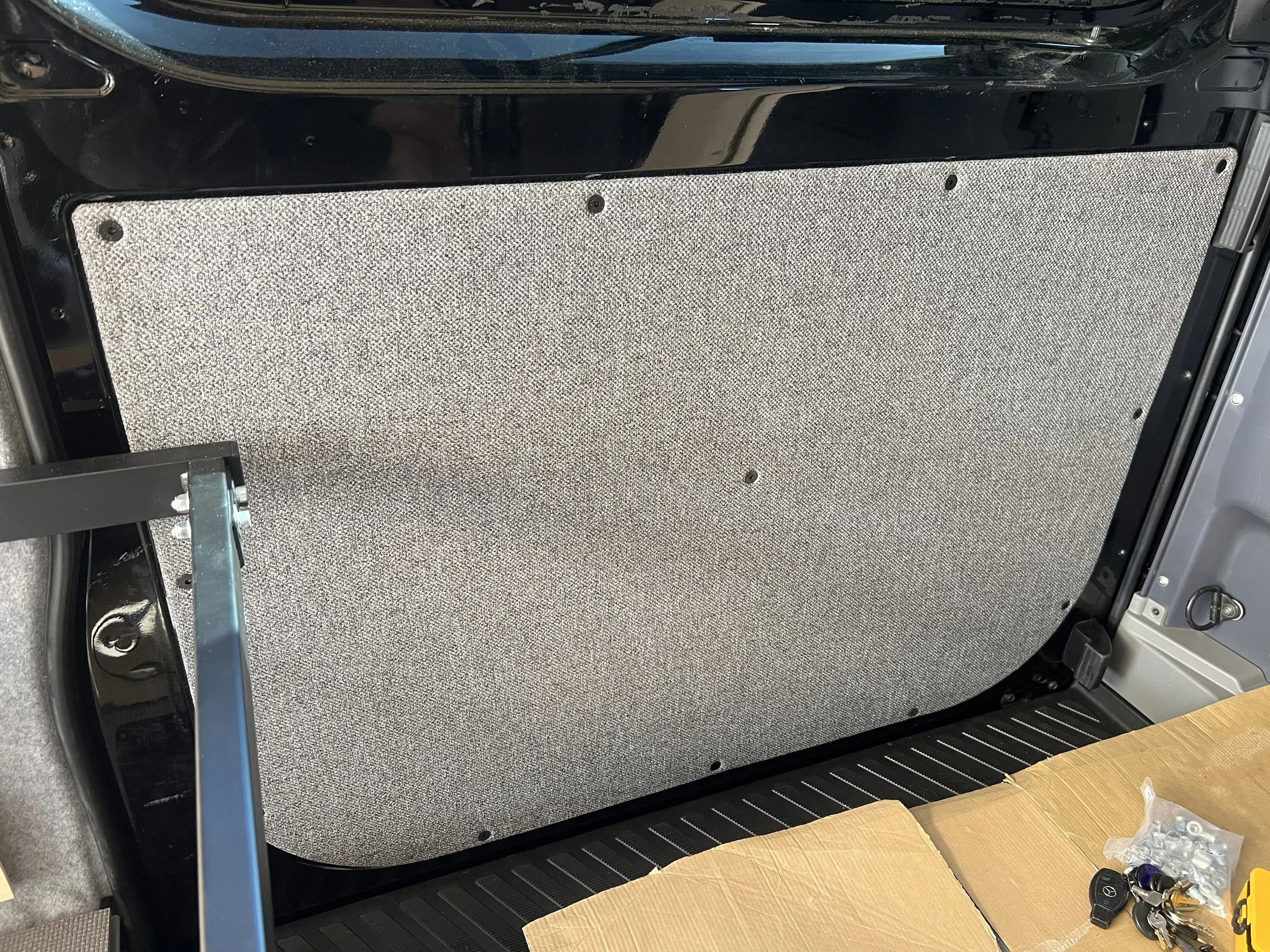 The interior of a vehicle showing a panel with a textured surface, screws, and some hardware items including keys and small tools on a cardboard box.