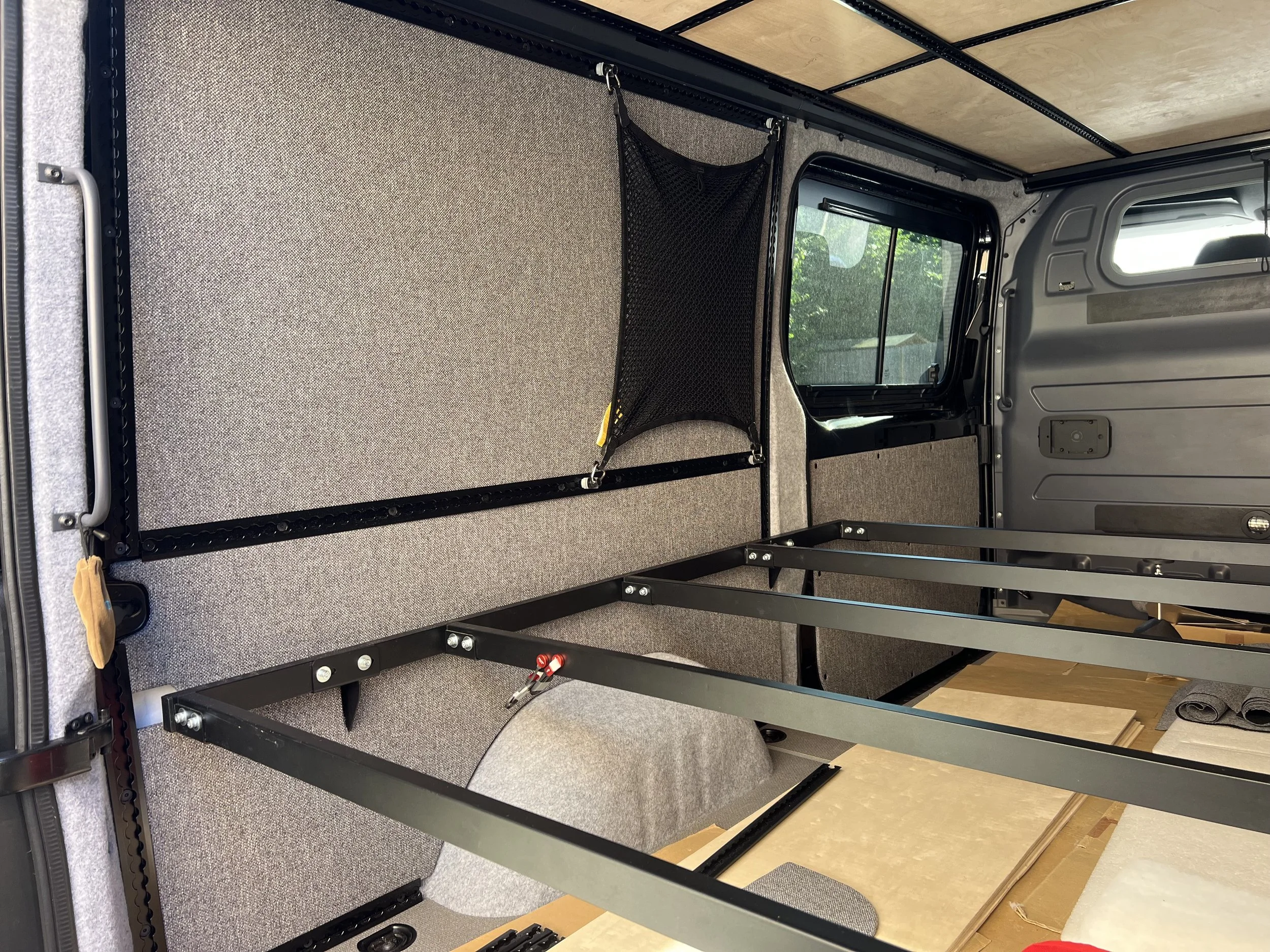 Interior of a vehicle being converted, with a partially installed bed frame and storage net on the wall, gray fabric wall panels, and construction materials like wood and fabric on the floor.