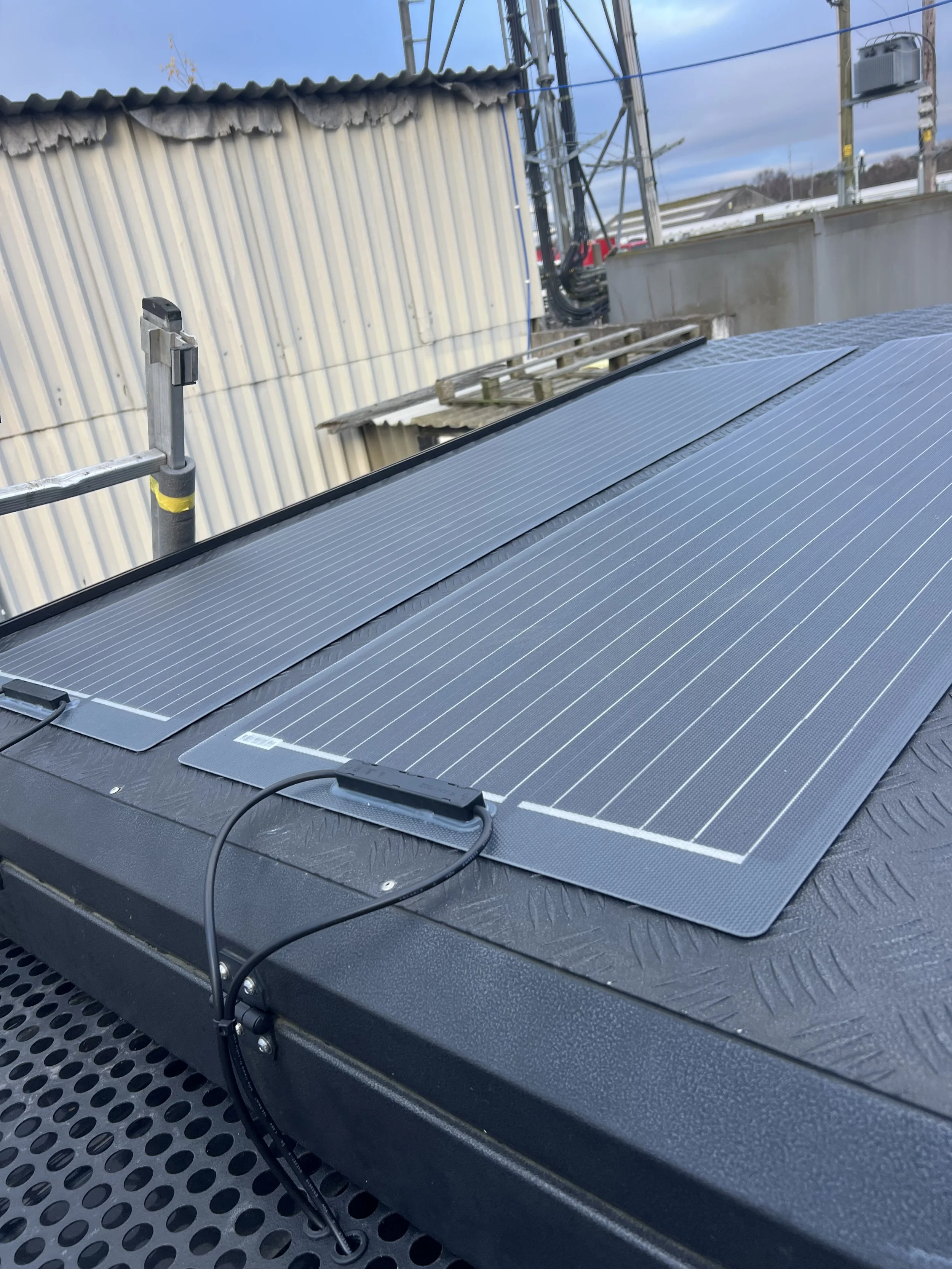 Close-up of a solar panel installed on a rooftop, with electrical wiring connected to it. In the background, there are other rooftops, utility poles, and electrical towers under a partly cloudy sky.