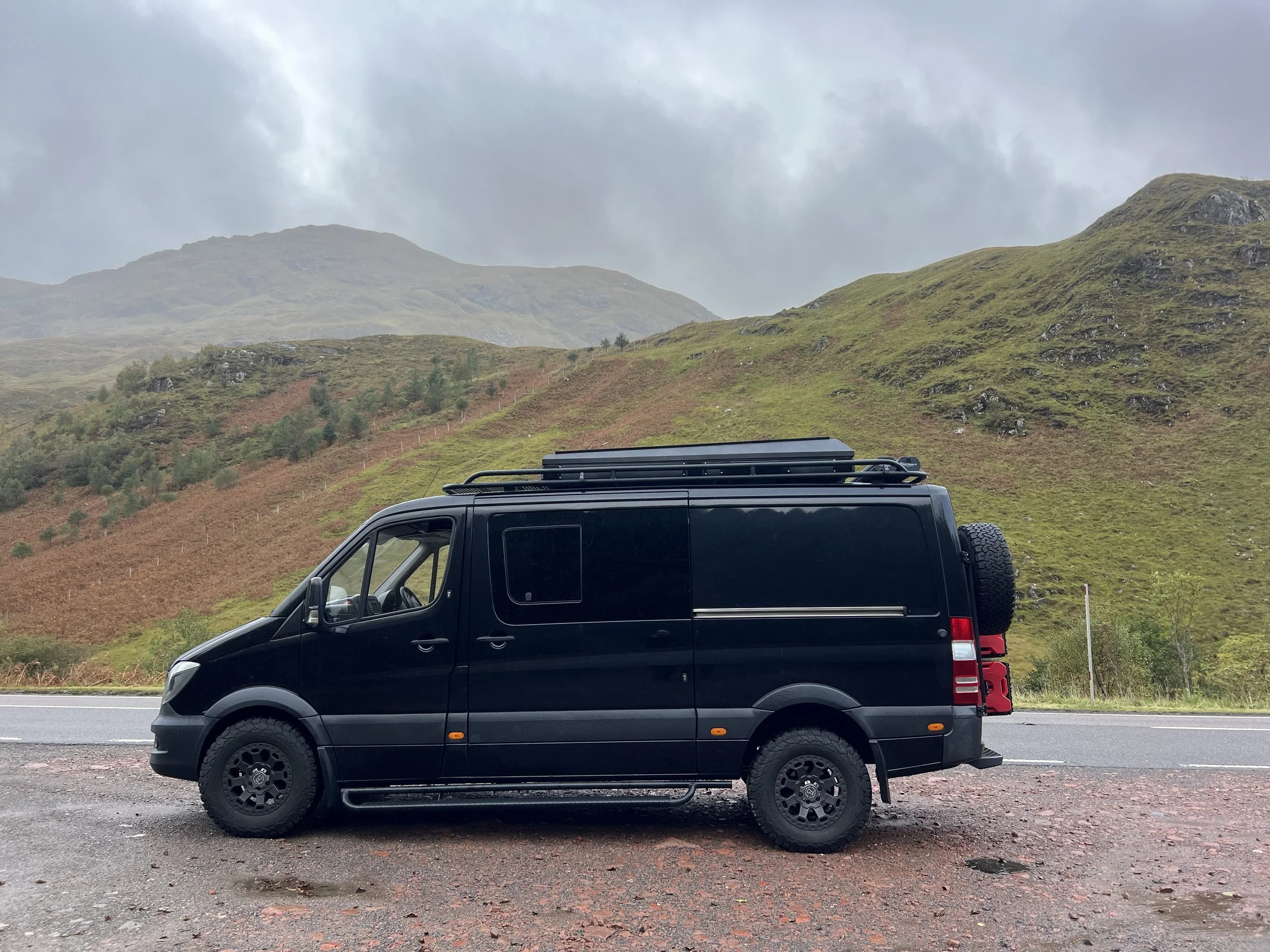 Black van parked on the side of a rural road with green hills and mountains in the background under cloudy skies.