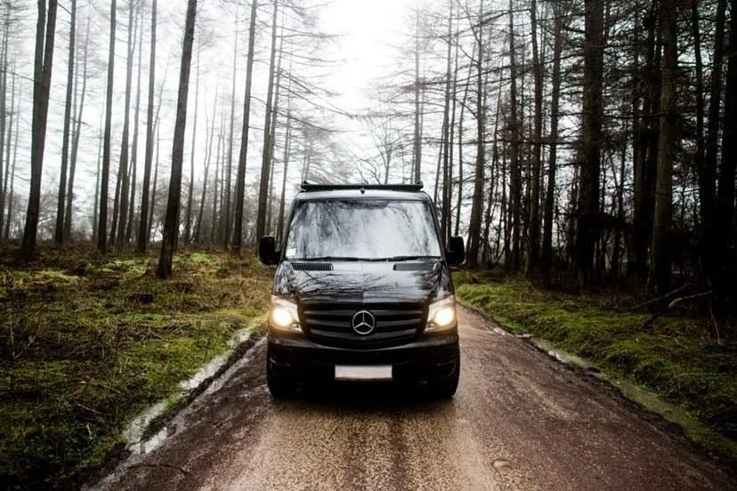 Black Mercedes-Benz van driving on a narrow dirt road through a wooded area with tall, leafless trees and mossy ground, overcast sky.