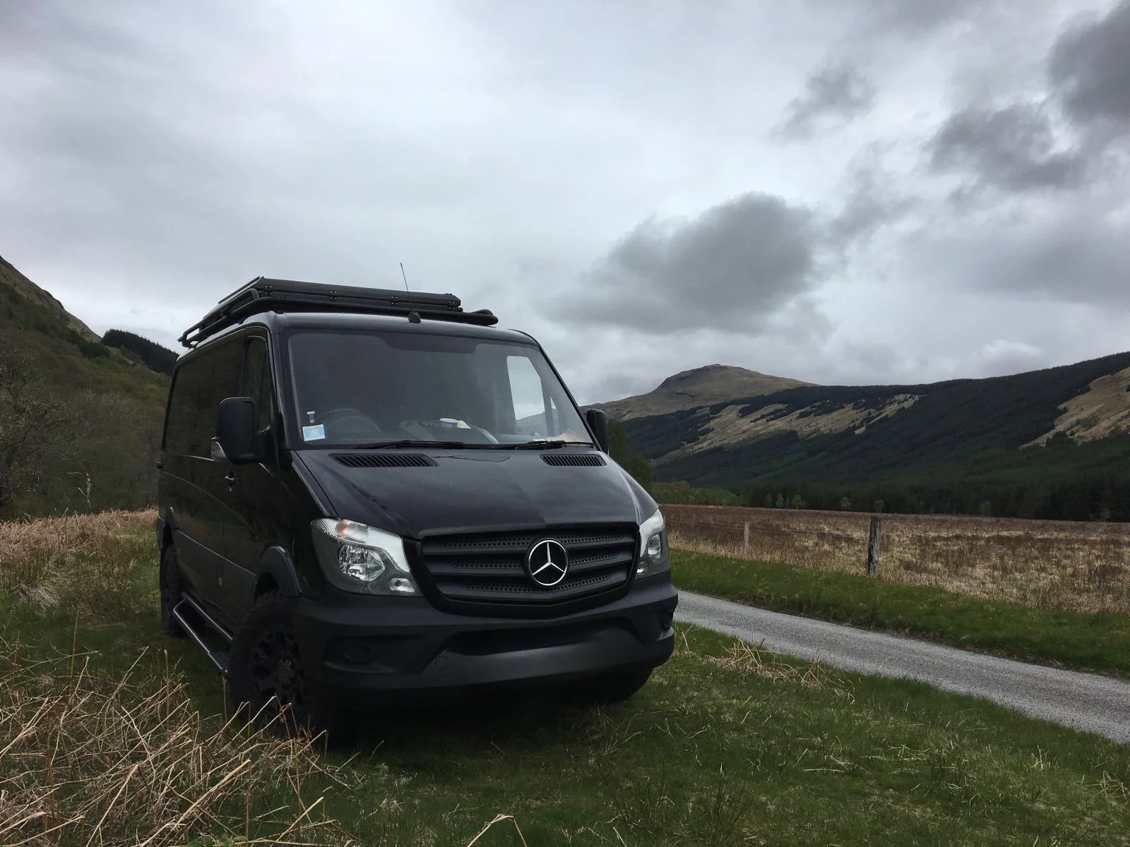 Black Mercedes-Benz van parked beside a rural road with mountains in the background under cloudy skies.