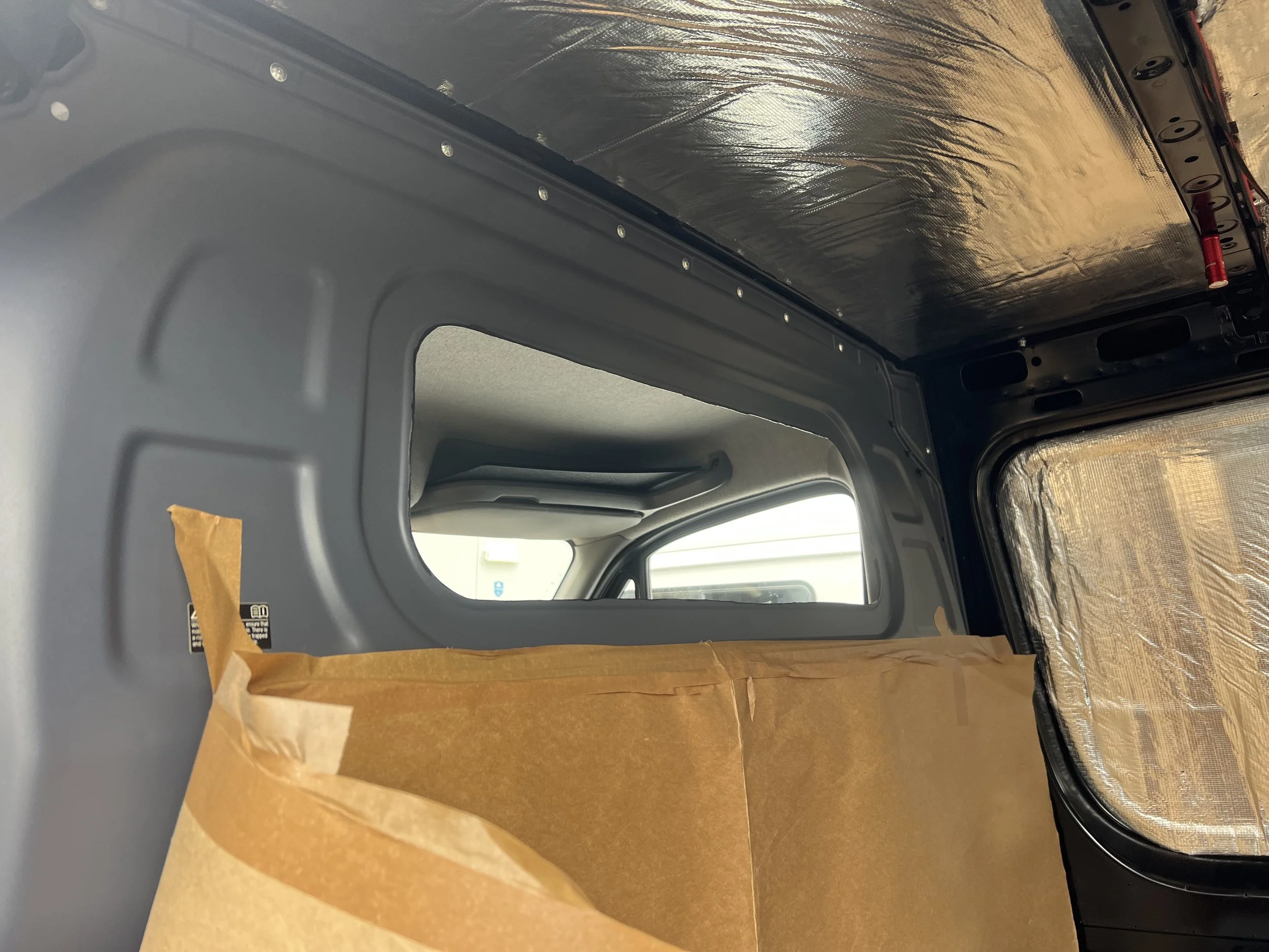 Interior of a vehicle with insulation material on the roof and window, and a cardboard piece taped to the side.