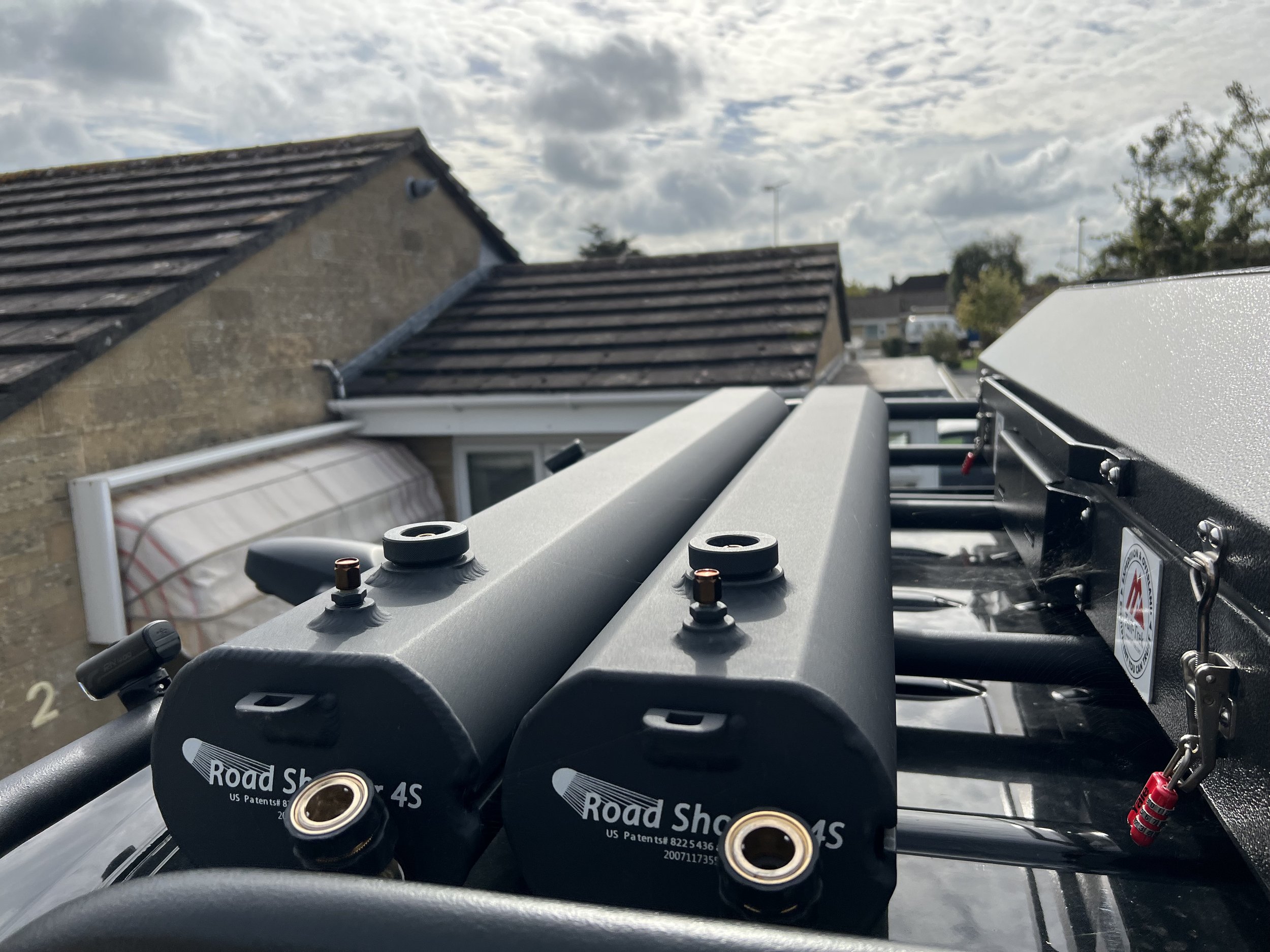 Close-up of two black cylindrical devices labeled 'Road Shock 4S' mounted on the roof of a vehicle in a residential neighborhood, with houses and cloudy sky in the background.