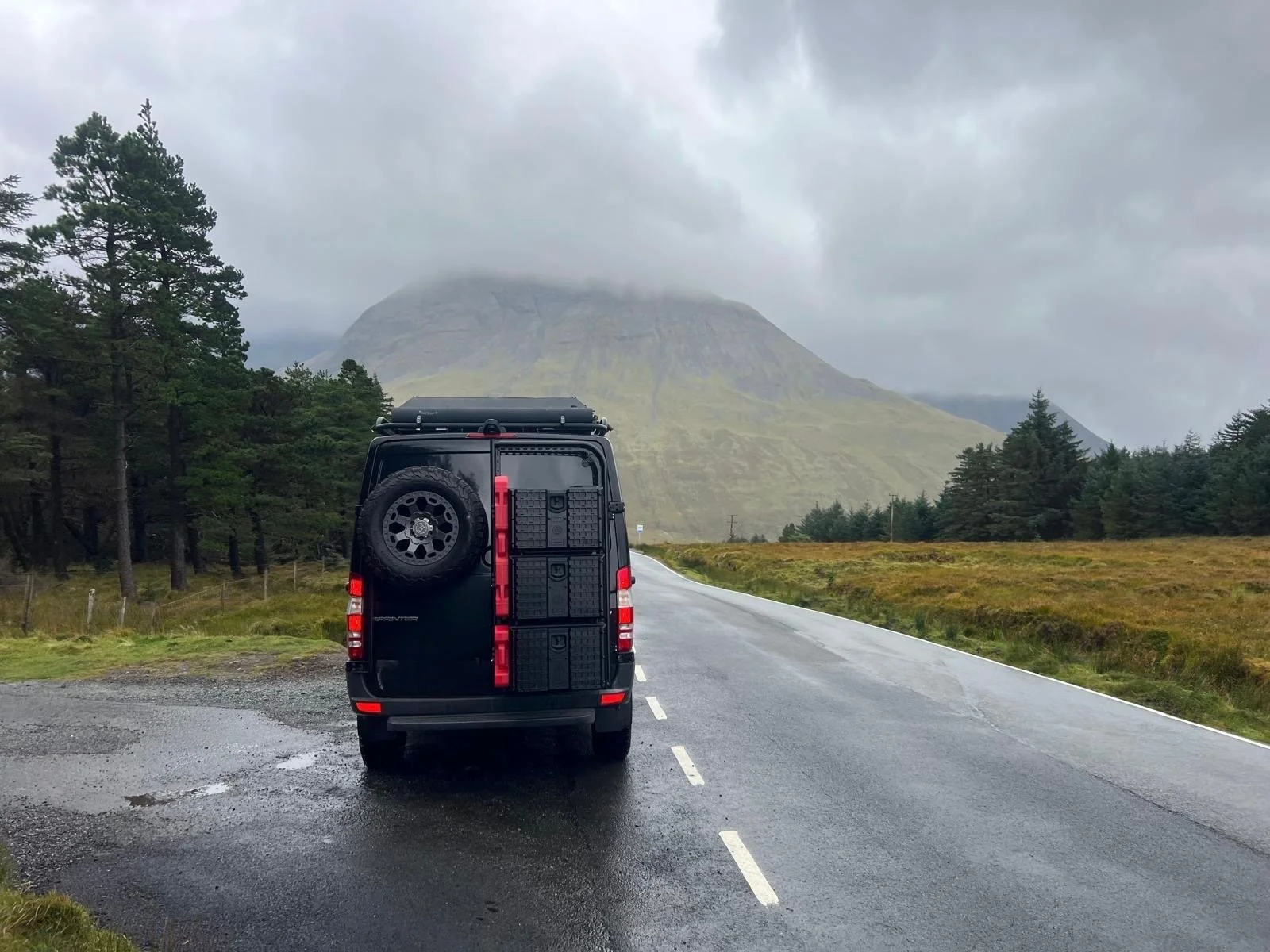 A black van parked on the side of a wet, empty road with a mountainous landscape in the background under cloudy skies.
