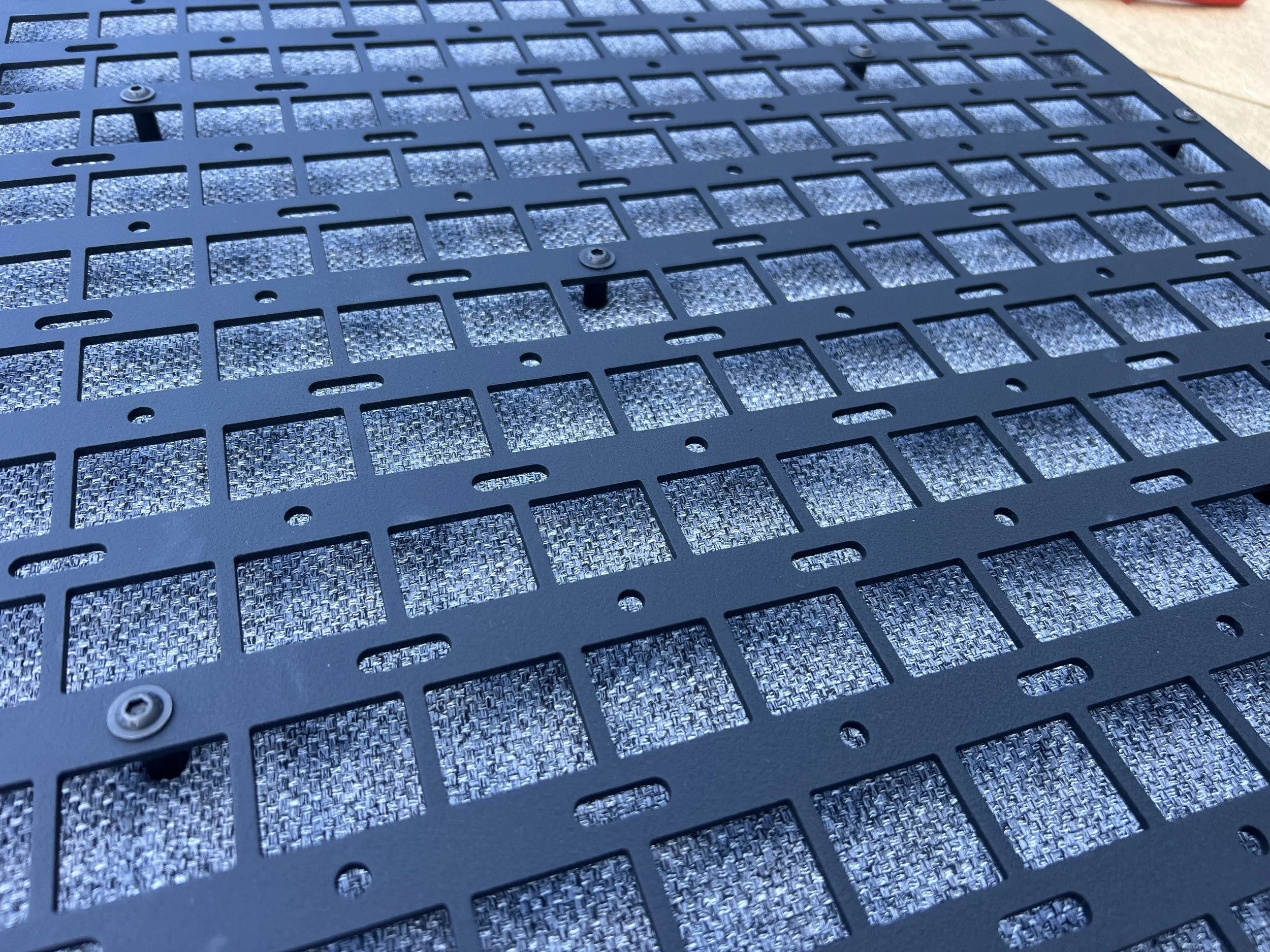 Close-up of a black metal grill or panel with rectangular cutouts, screws, and a textured surface underneath.