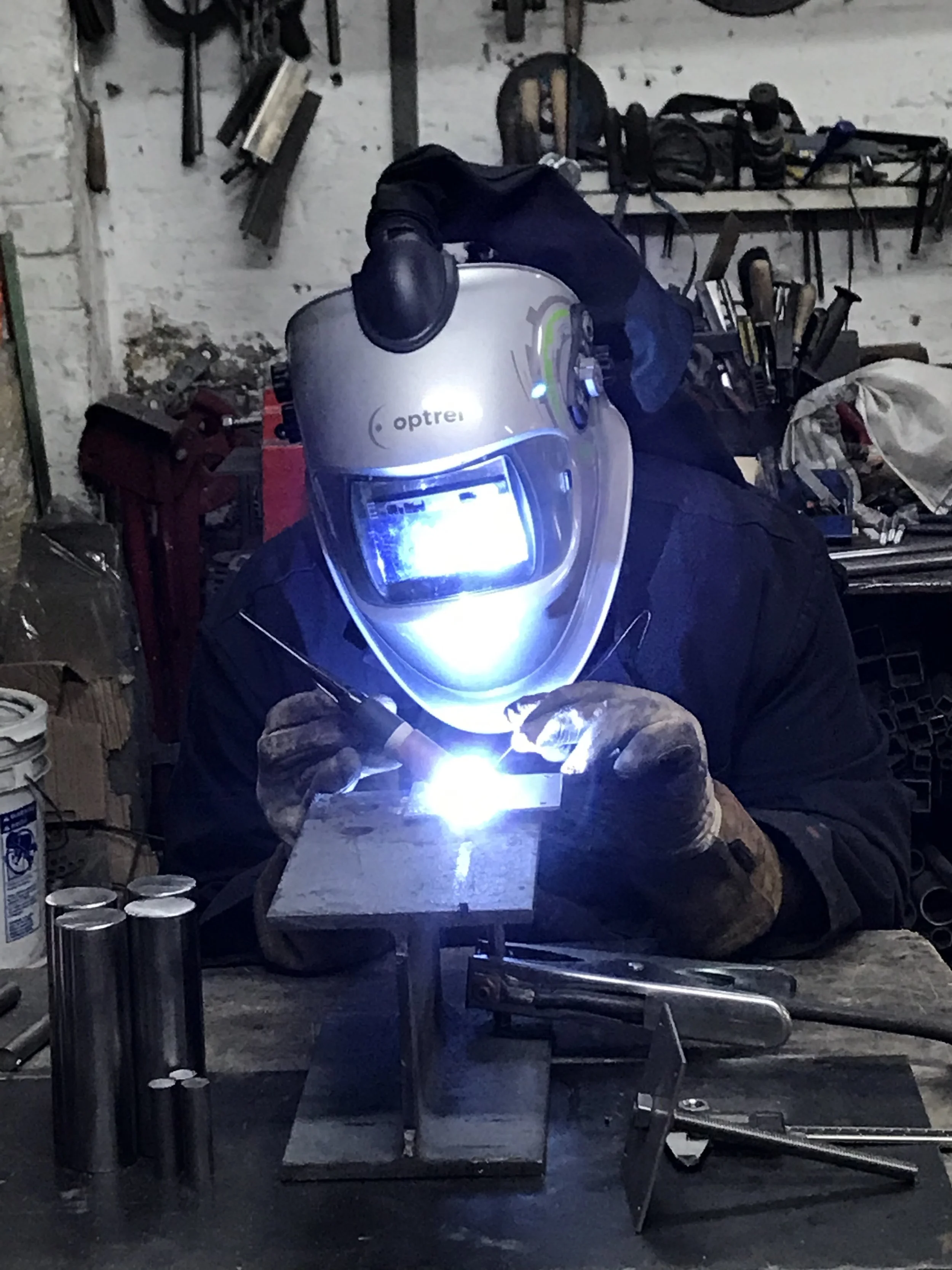 A person welding metal on a workbench, wearing a protective welding helmet and gloves, in a workshop with tools and equipment in the background.