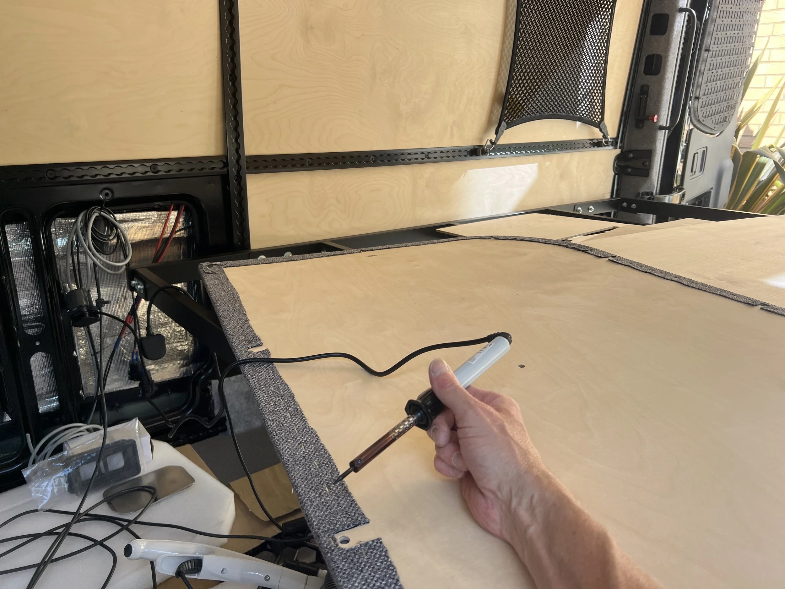 A person is holding a soldering iron and working on electronic components on a wooden surface, with various wires and tools nearby.