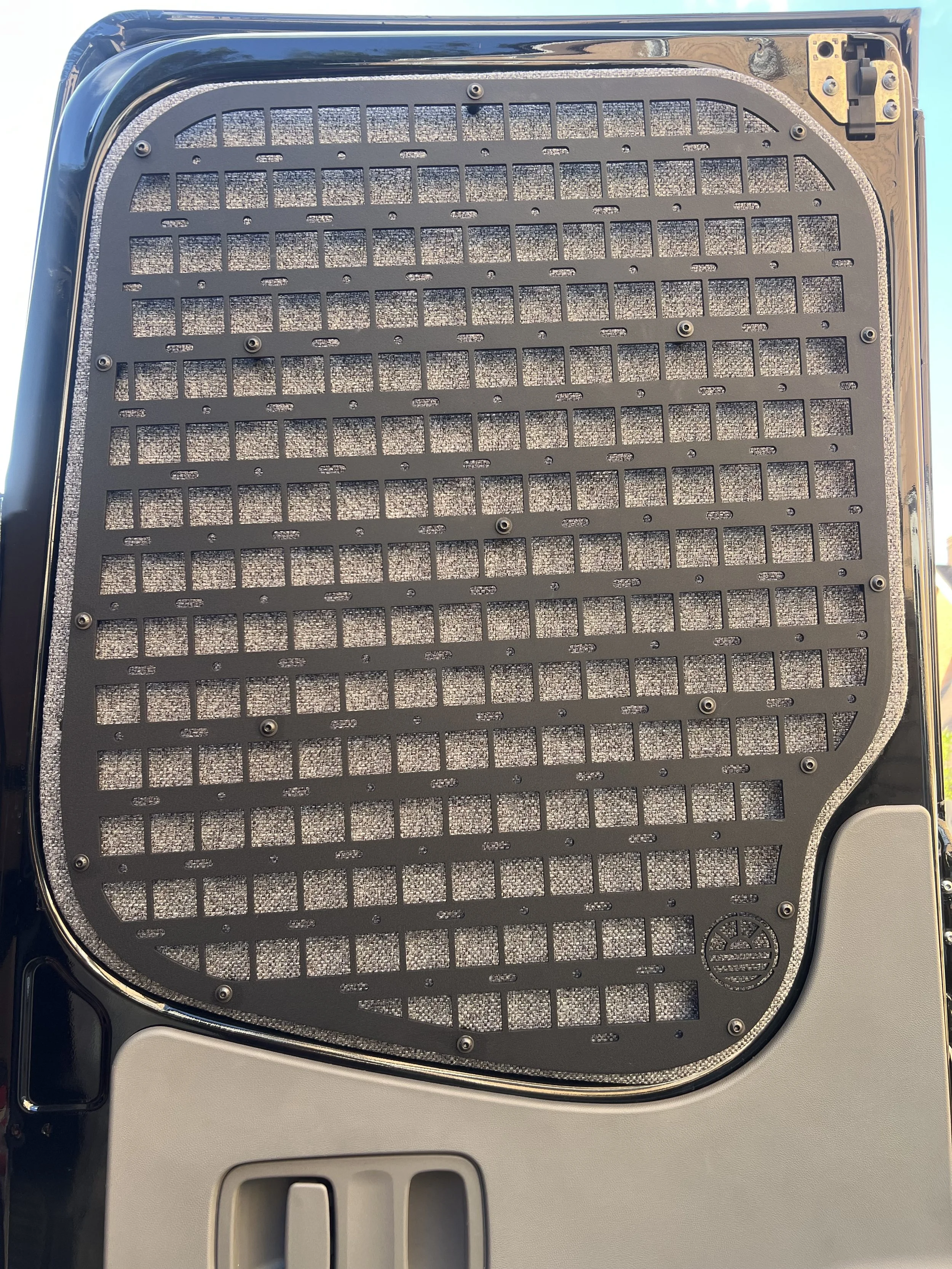 Close-up view of a black, rectangular grill with small square openings, mounted on an outdoor appliance or device, with a rounded corner on the top right and a gray vent at the bottom.