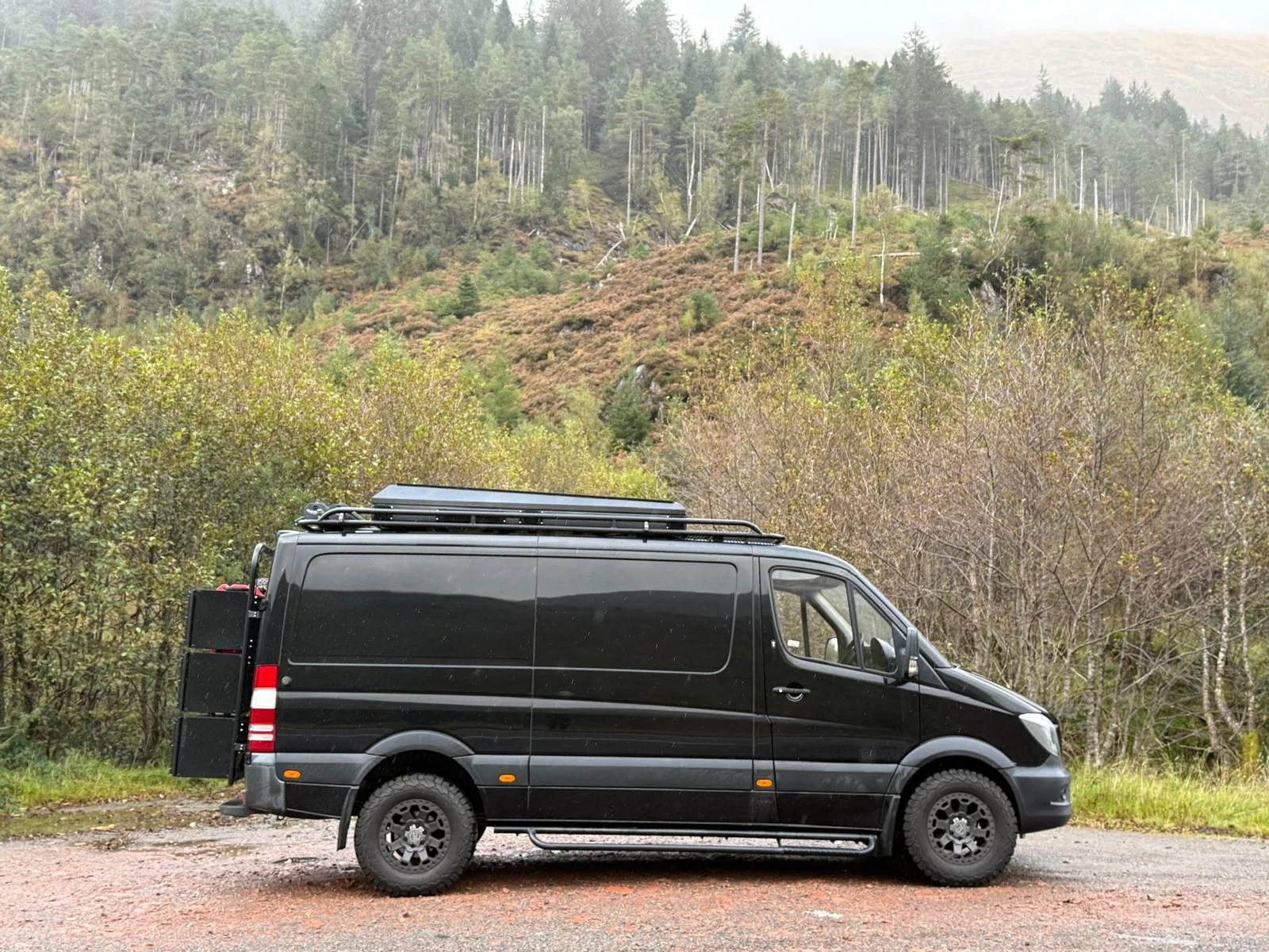 A black cargo van parked on a gravel surface with trees and hills in the background.