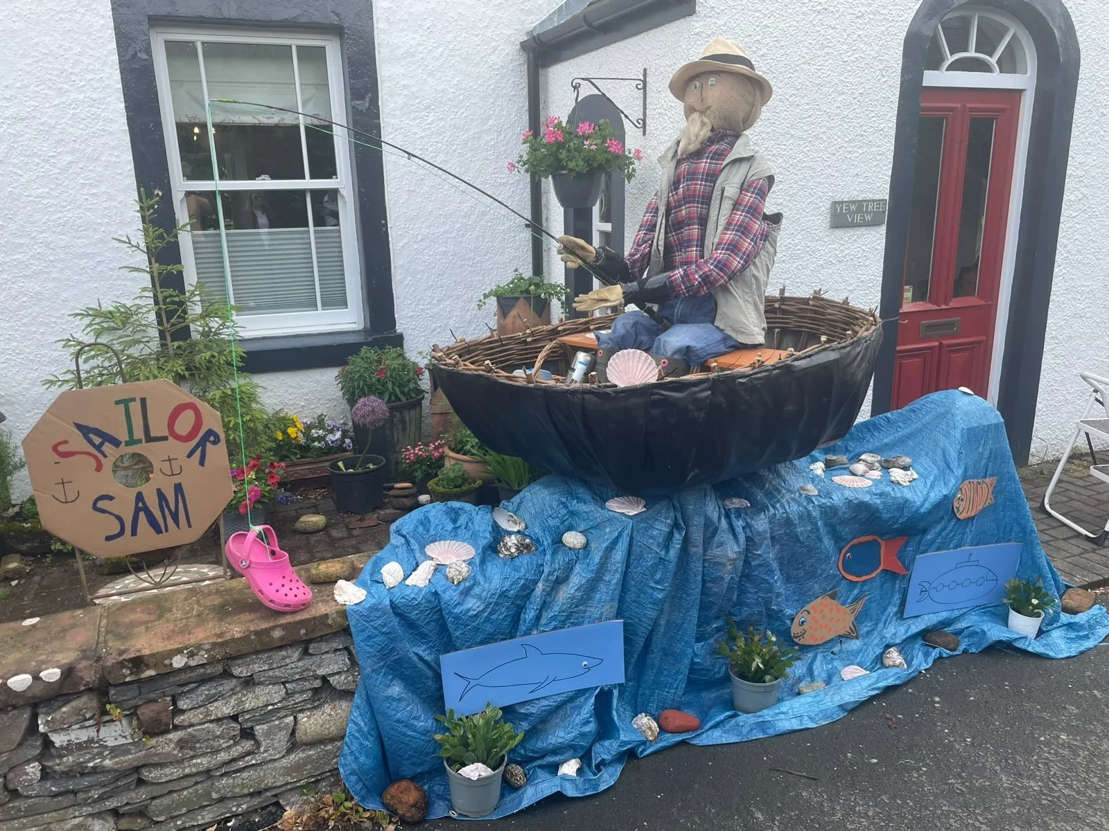 A garden display featuring a scarecrow fishing from a boat, with a blue cloth decorated with drawings of fish, shells, and rocks. A sign reads 'Sailor Sam.' There are potted plants and flowers, shells, and seashell-themed decorations around the displ