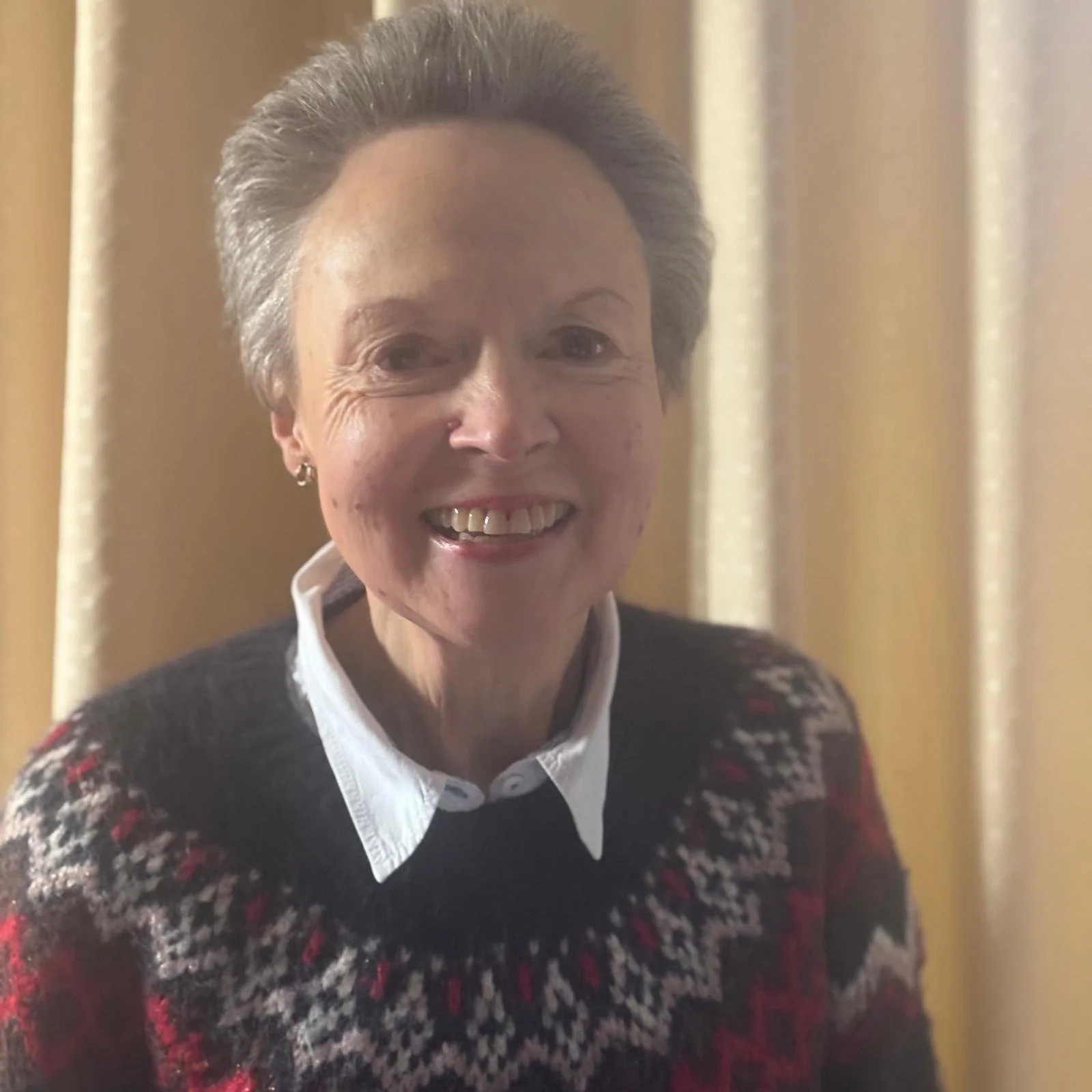 A smiling elderly woman with short gray hair, wearing a patterned sweater over a white collared shirt, standing in front of beige curtains.
