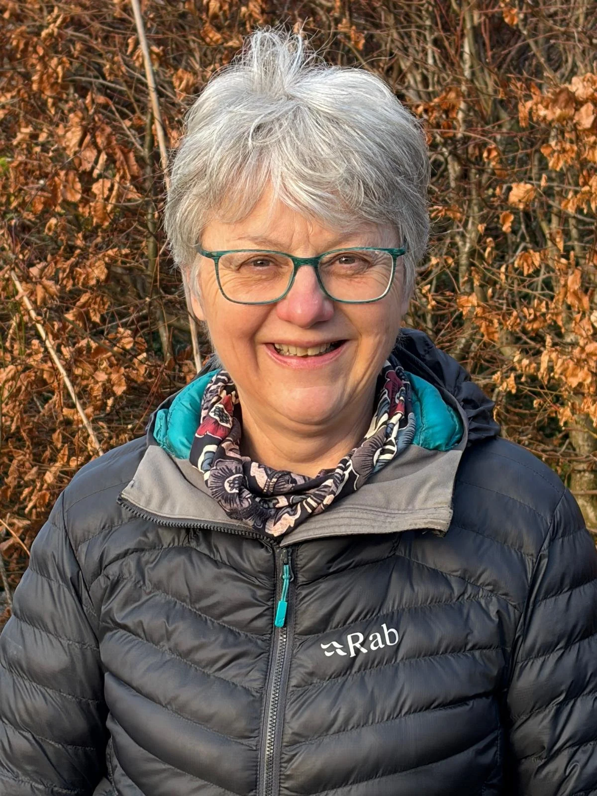 Close-up of smiling elderly woman with gray hair and glasses, wearing a black Rab jacket and a patterned scarf, standing outdoors in front of brown foliage.