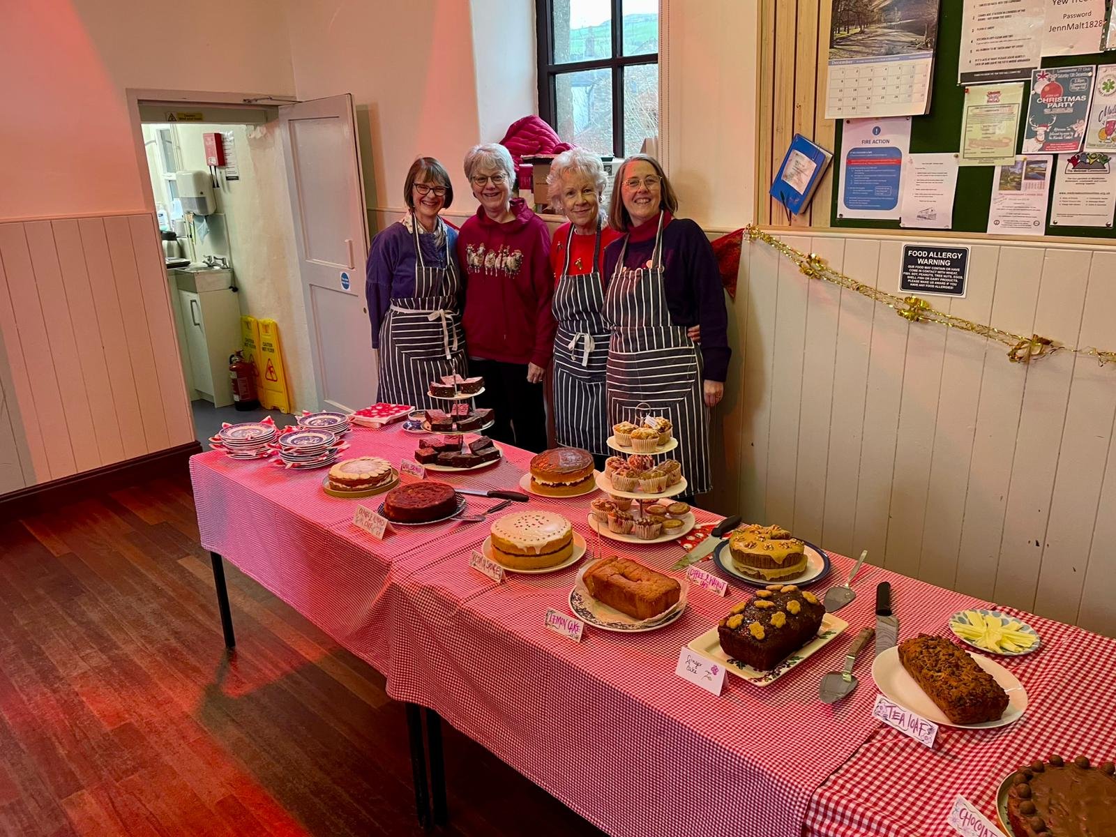 Four women standing behind a table with various homemade cakes and baked goods, in a room with wooden walls and a window.