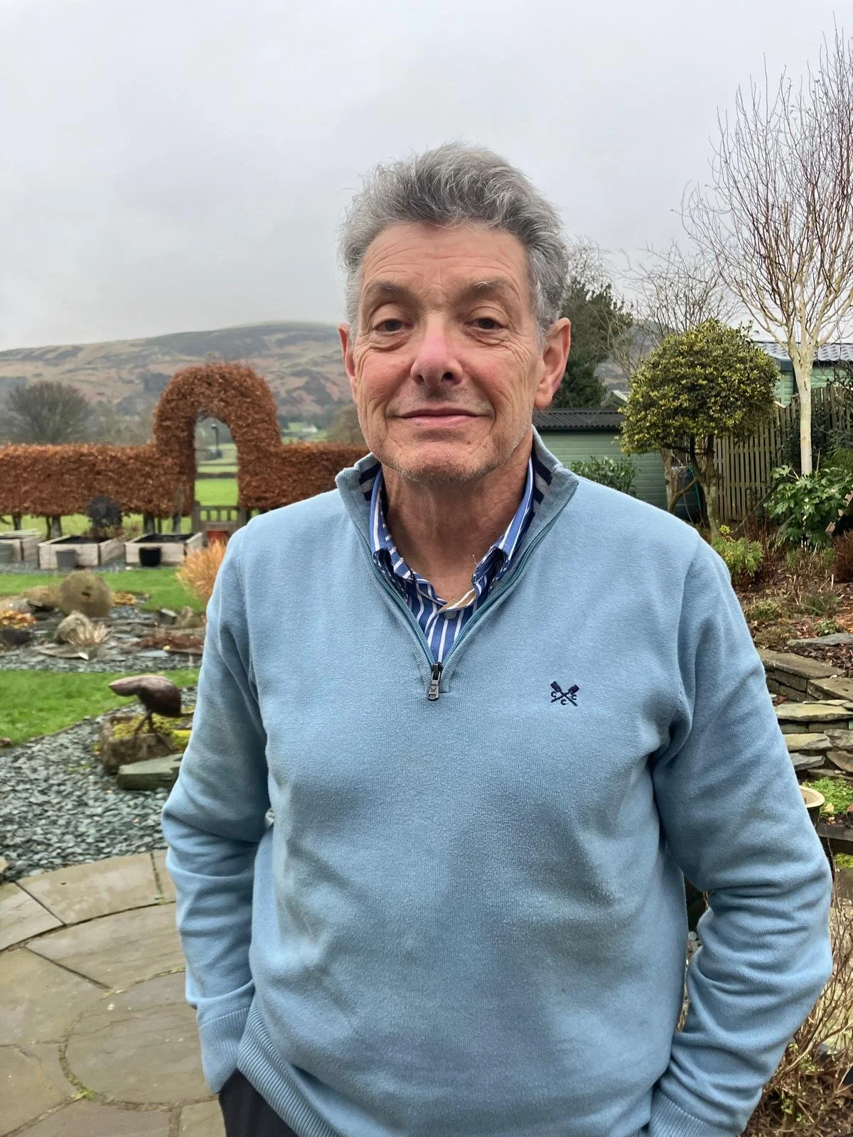 A middle-aged man with gray hair standing outdoors in a garden with a mountainous background, wearing a light blue zip-up sweater over a striped shirt.