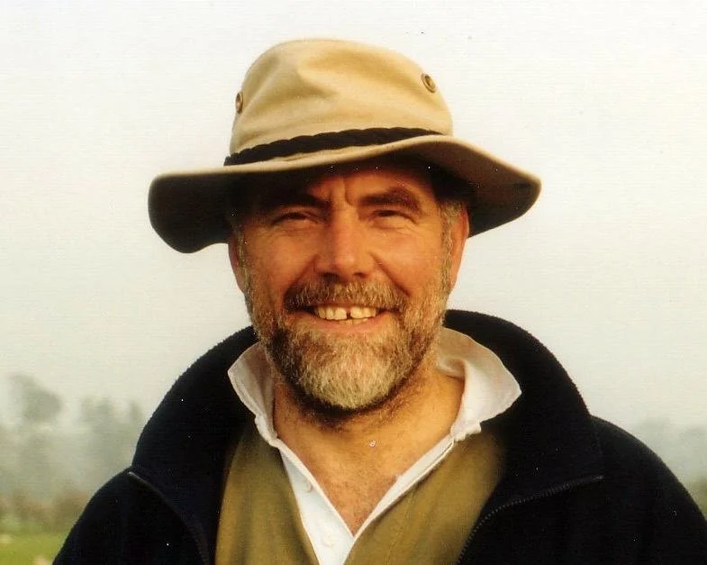 A man with a beard and mustache smiling, wearing a wide-brimmed beige hat and a dark jacket outdoors.