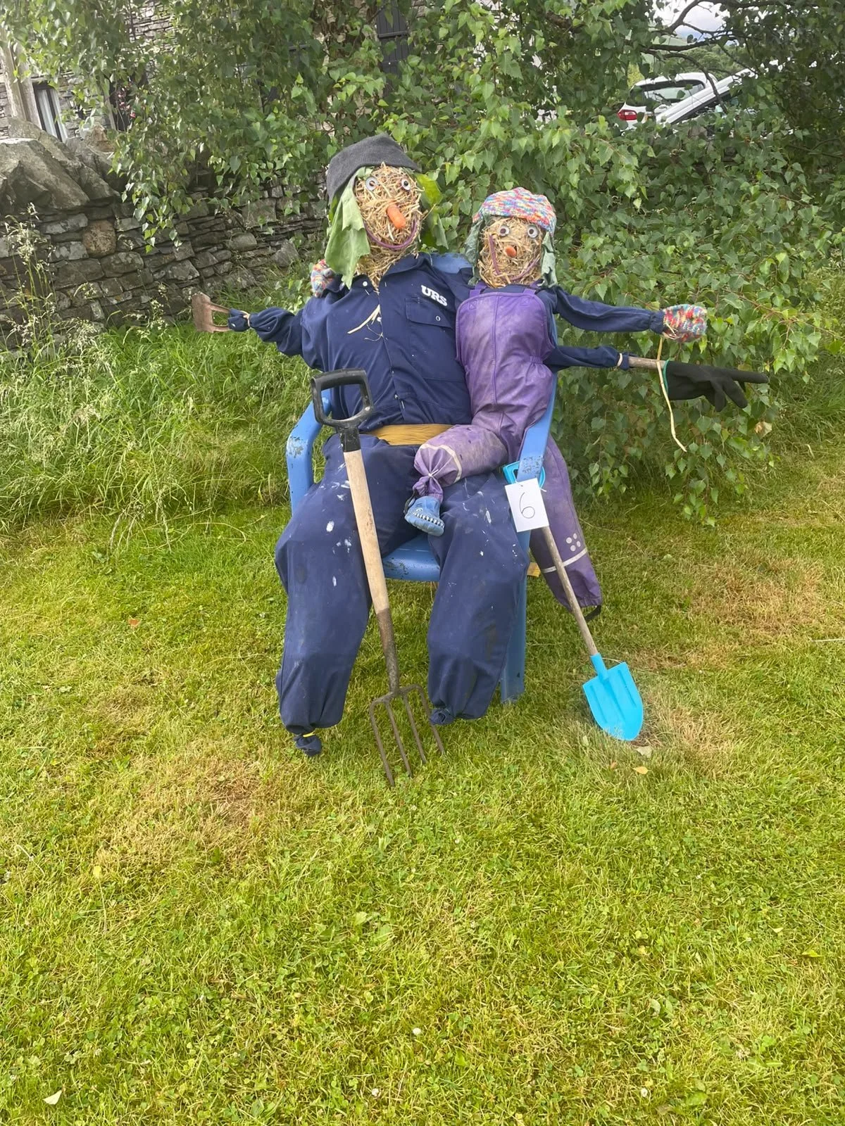 Decorative scarecrows resembling an older couple sitting on a blue plastic chair outside on grass, with gardening tools, in front of bushes and a stone wall.
