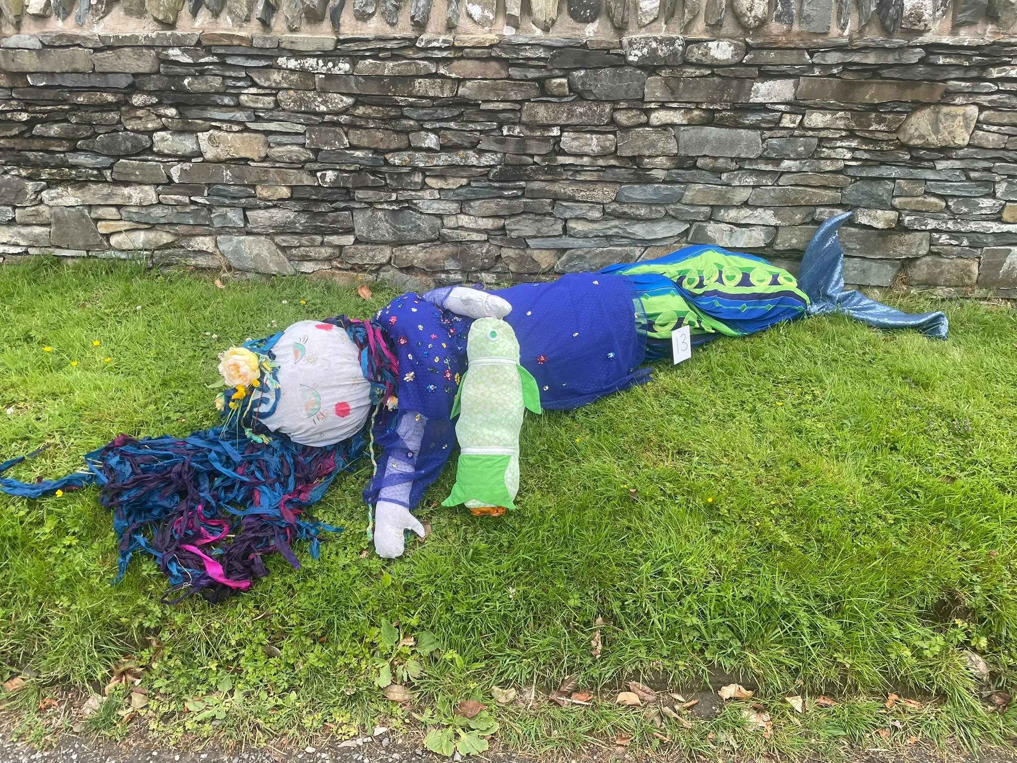 A green grass lawn with a stone wall in the background. On the grass, there are various stuffed and fabric items, including a pillow with a pink print, a stuffed animal, and several colorful fabric pieces, some of which are tied or scrunched.