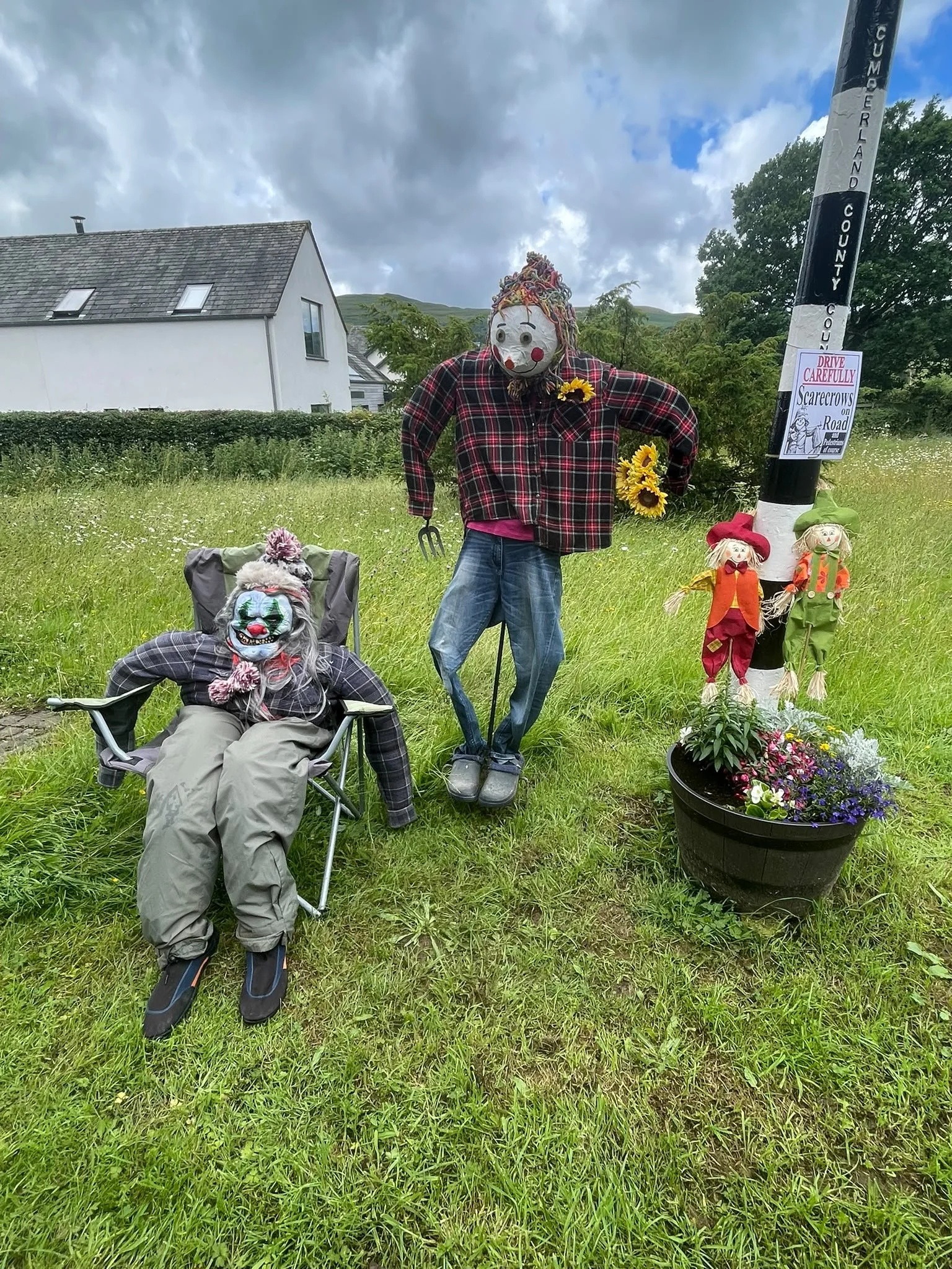 A scarecrow decorated with a clown mask, plaid shirt, and blue jeans, standing among sunflowers. Nearby are two smaller scarecrows and a large pot of colorful flowers, with a white house and cloudy sky in the background.