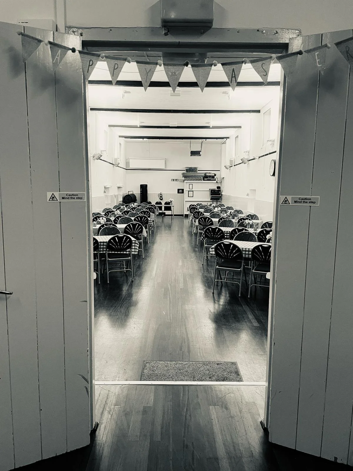 An entryway into a large room with neatly arranged tables and chairs, likely a community hall or event space, with a banner that says 'PARTY' hanging overhead.