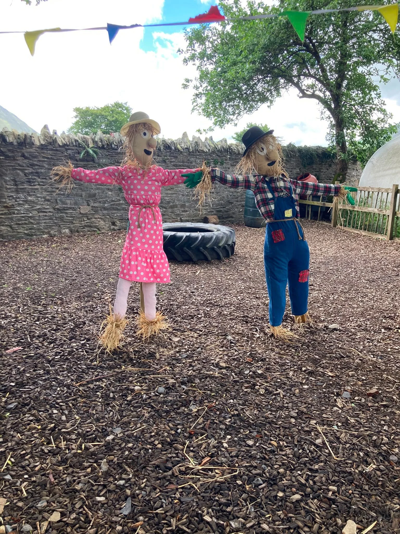 Two scarecrows in a yard, holding hands. One is dressed in pink polka-dot dress with a straw hat, and the other in plaid shirt and blue overalls with a black hat. The scarecrows have straw hair and expressive faces, with a tire, barrels, and a stone 