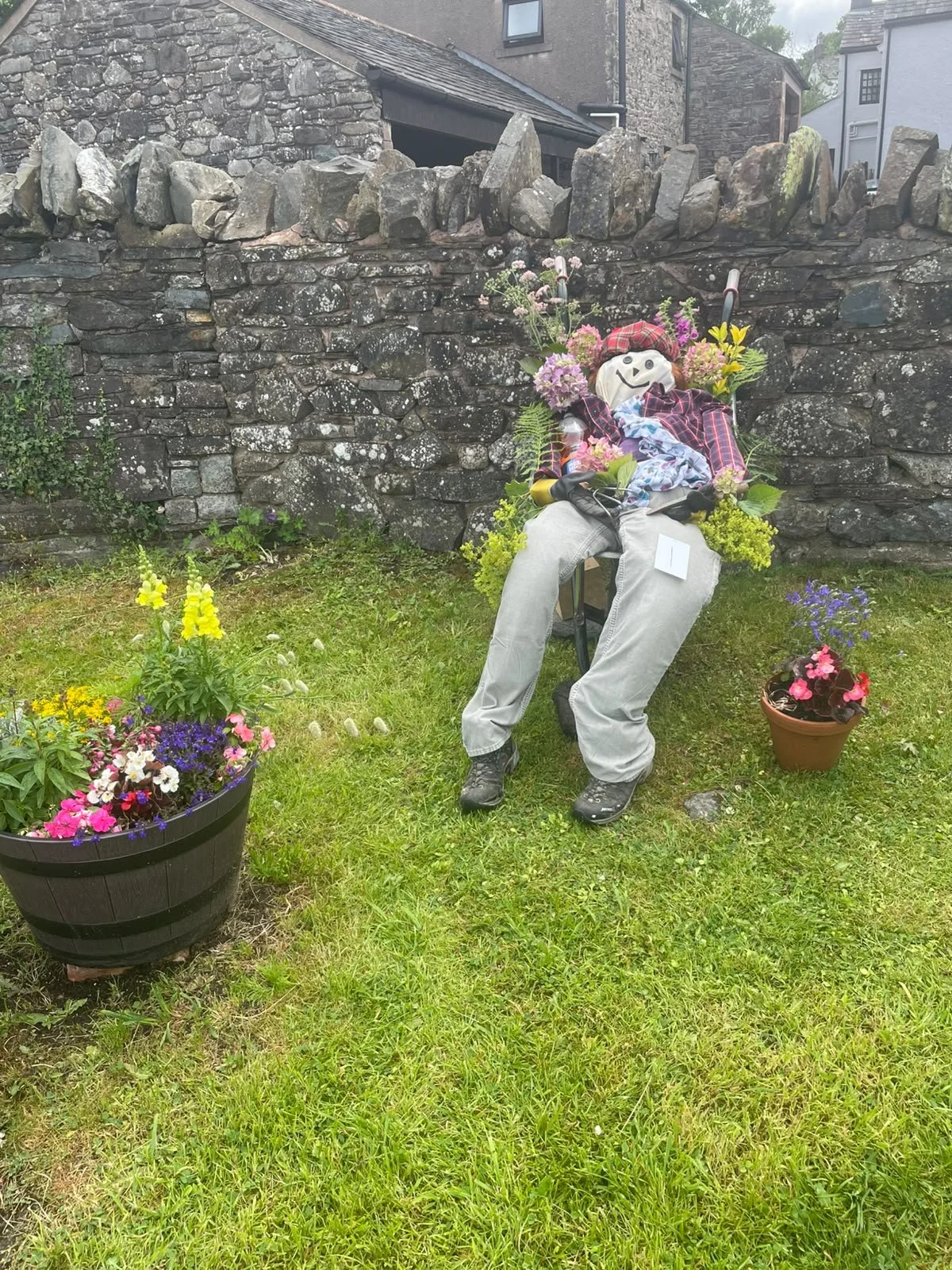 A scarecrow-like figure with a smiley face, wearing a red checkered shirt, beige pants, and a red hat, seated on a chair in a garden with colorful flowers and a stone wall in the background.