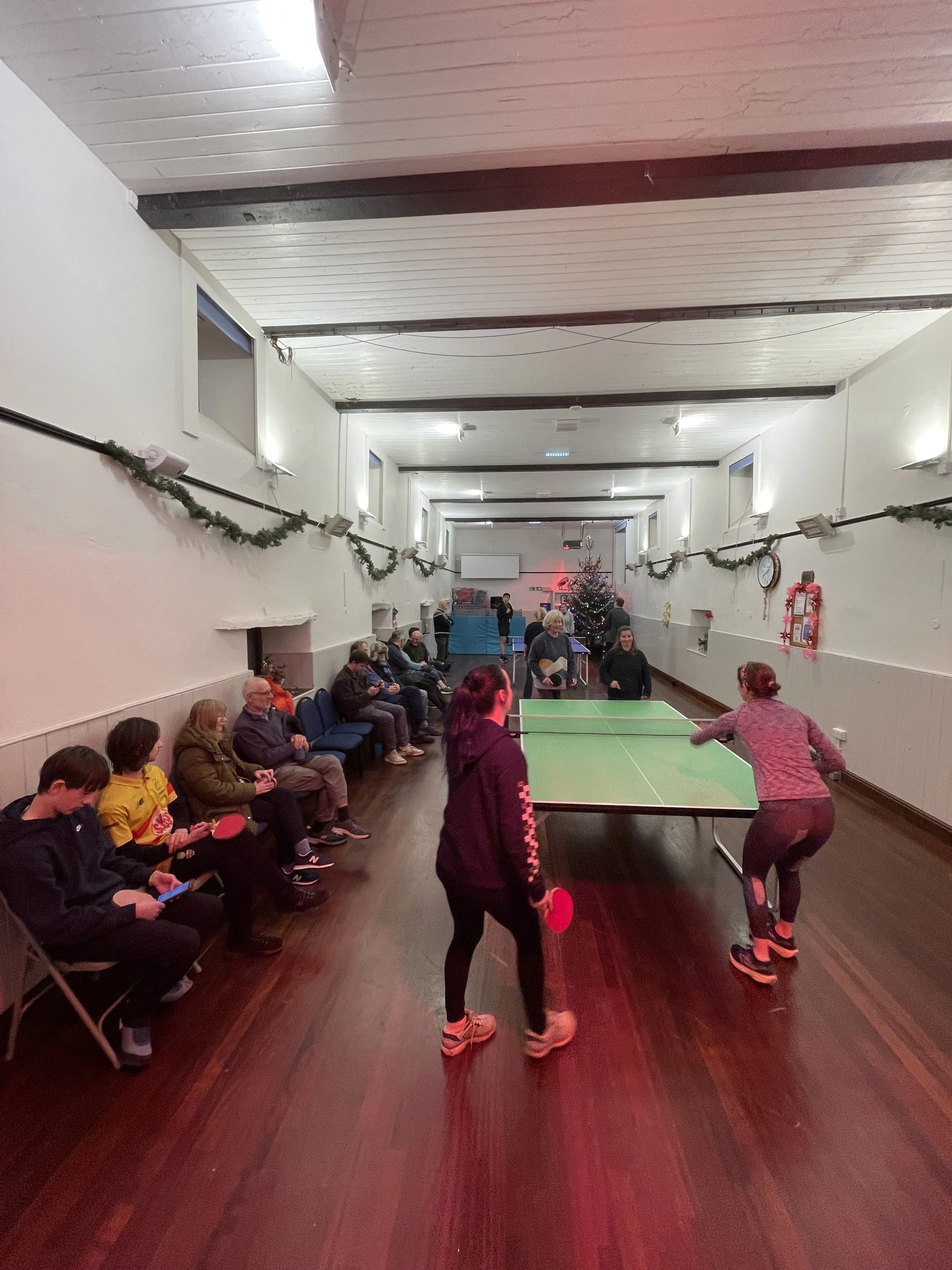Indoor room with people watching and playing ping pong near a Christmas tree, decorated for the holidays.