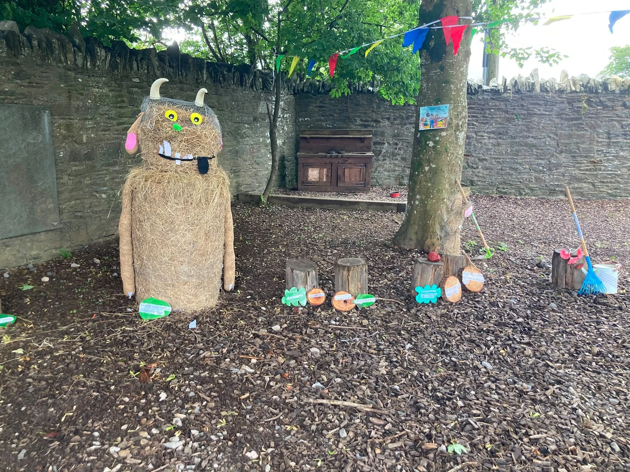 Decorative outdoor scene featuring a straw figure with cartoonish eyes, horns, and exaggerated facial features, surrounded by colorful banners, painted rocks with labels, and gardening tools, set against a background of a stone wall and trees.
