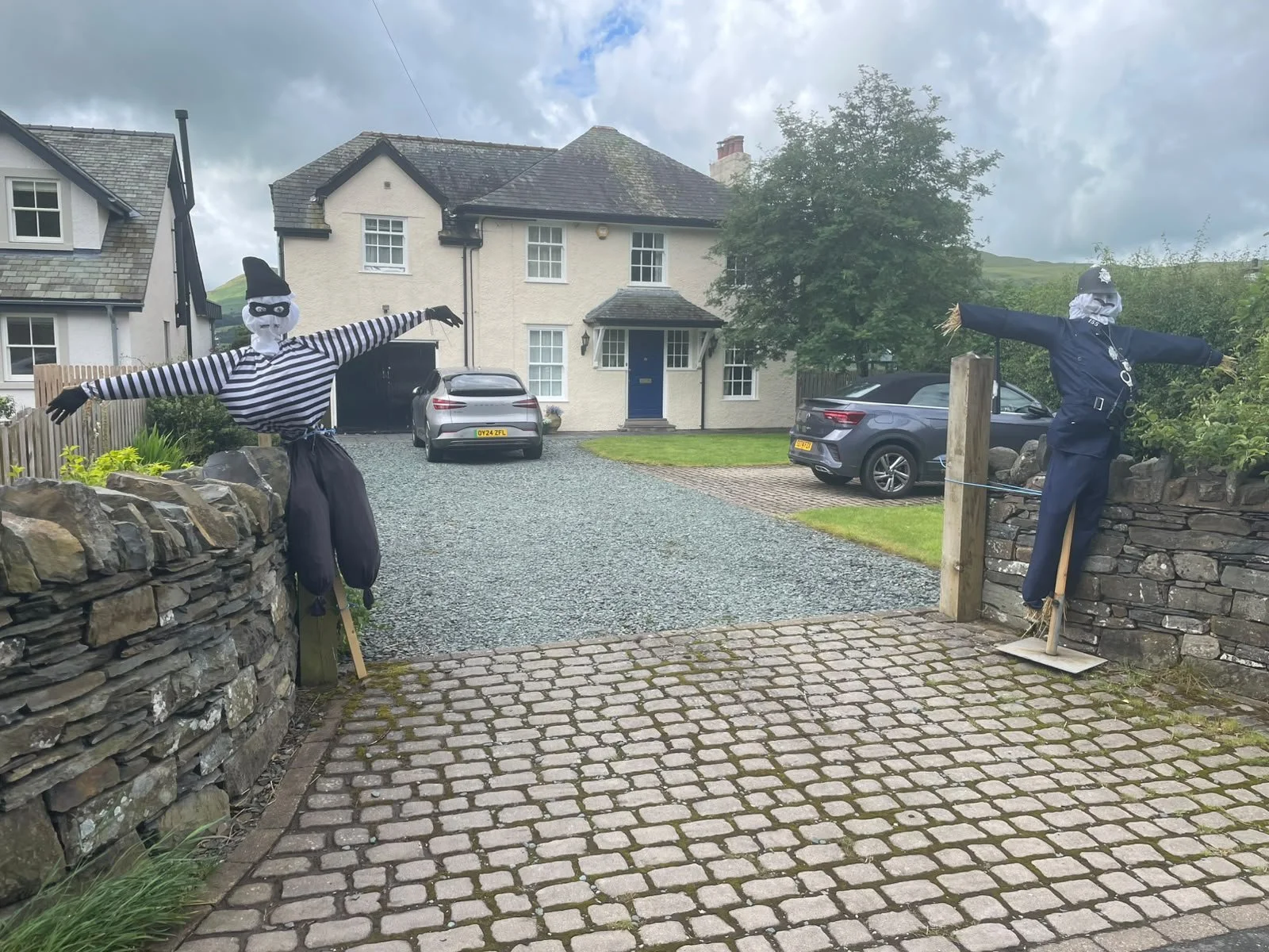 Two scarecrows dressed as burglars standing on opposite sides of a driveway, with a house in the background and cars parked outside. One scarecrow is wearing a striped shirt and a hat, and the other is in a police uniform. The scene appears to be a d