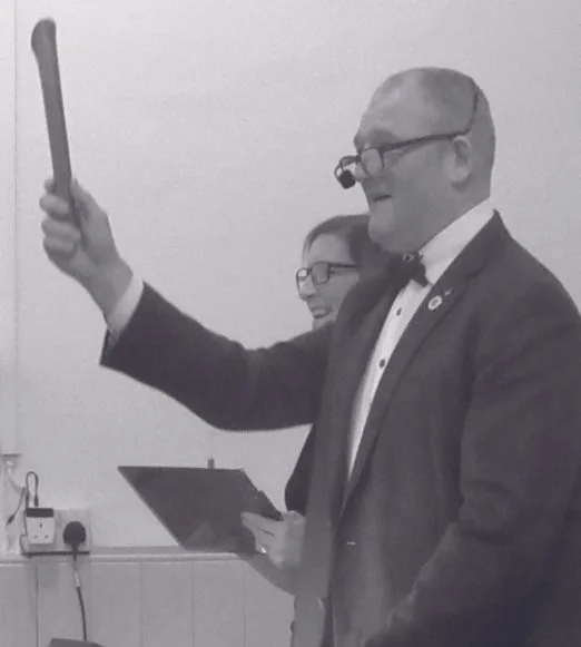 A man in formal attire, wearing glasses and a bow tie, is smiling and holding a wooden gavel or stick in his right hand. There is a woman in the background holding a clipboard, also smiling. They appear to be at an event or presentation.