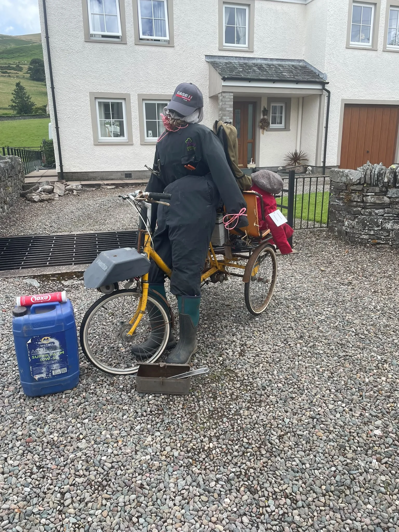 A person with a mask and hat standing next to a yellow bicycle with a rear basket, on a gravel driveway in front of a white house. Items on the ground include a blue container, a small metal box, and a rectangular object. The house has a wooden door,