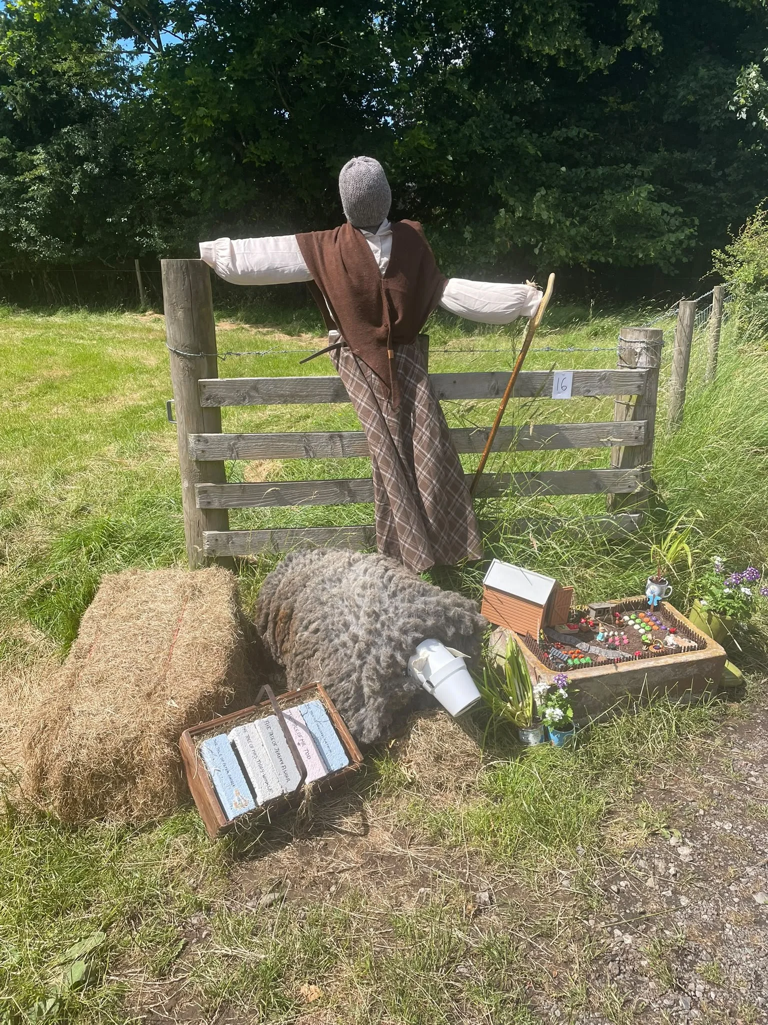 A scarecrow dressed in historical clothing, with outstretched arms, standing behind a wooden fence in a grassy field with trees in the background. In front of the scarecrow, there are hay bales, a sheep costume, and some small decorative items includ