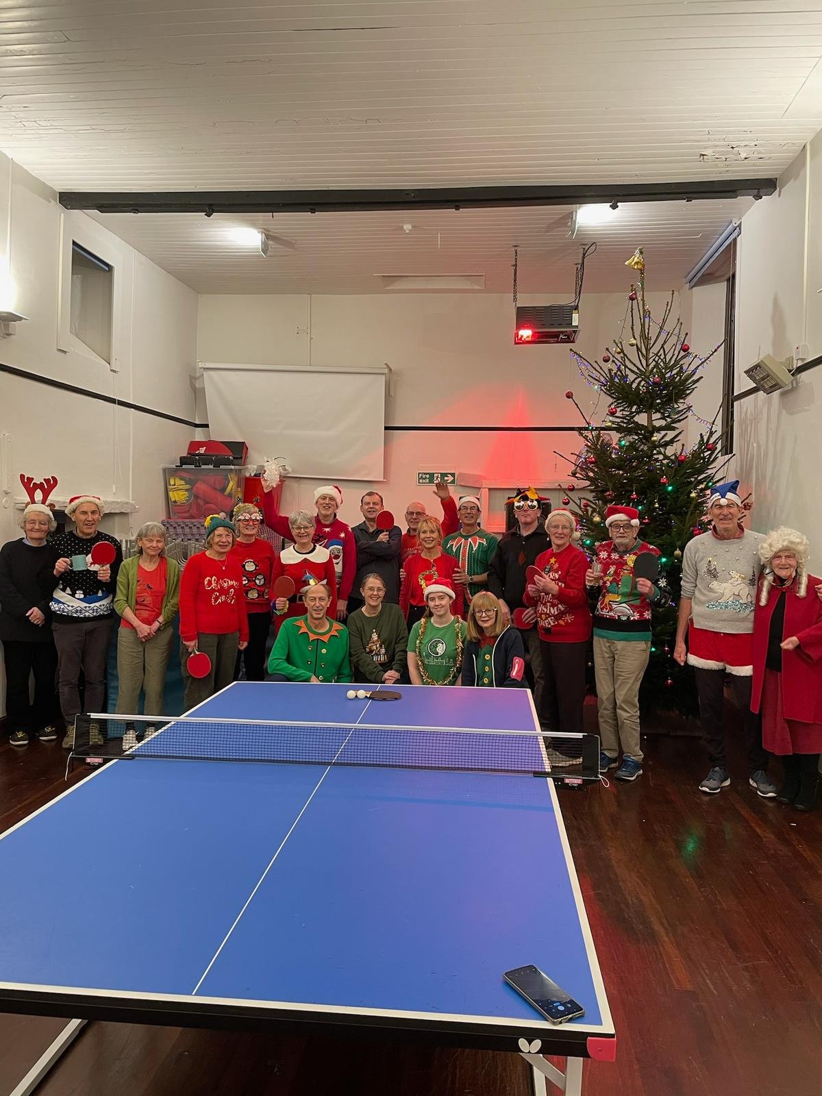 Group of people celebrating Christmas with a decorated Christmas tree, some dressed in Santa hats, and playing table tennis.
