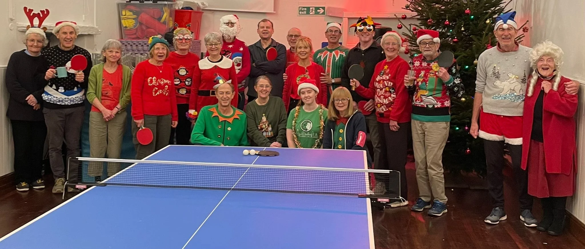 Group of people celebrating Christmas, many wearing festive sweaters and accessories, around a ping pong table with a Christmas tree in the background.