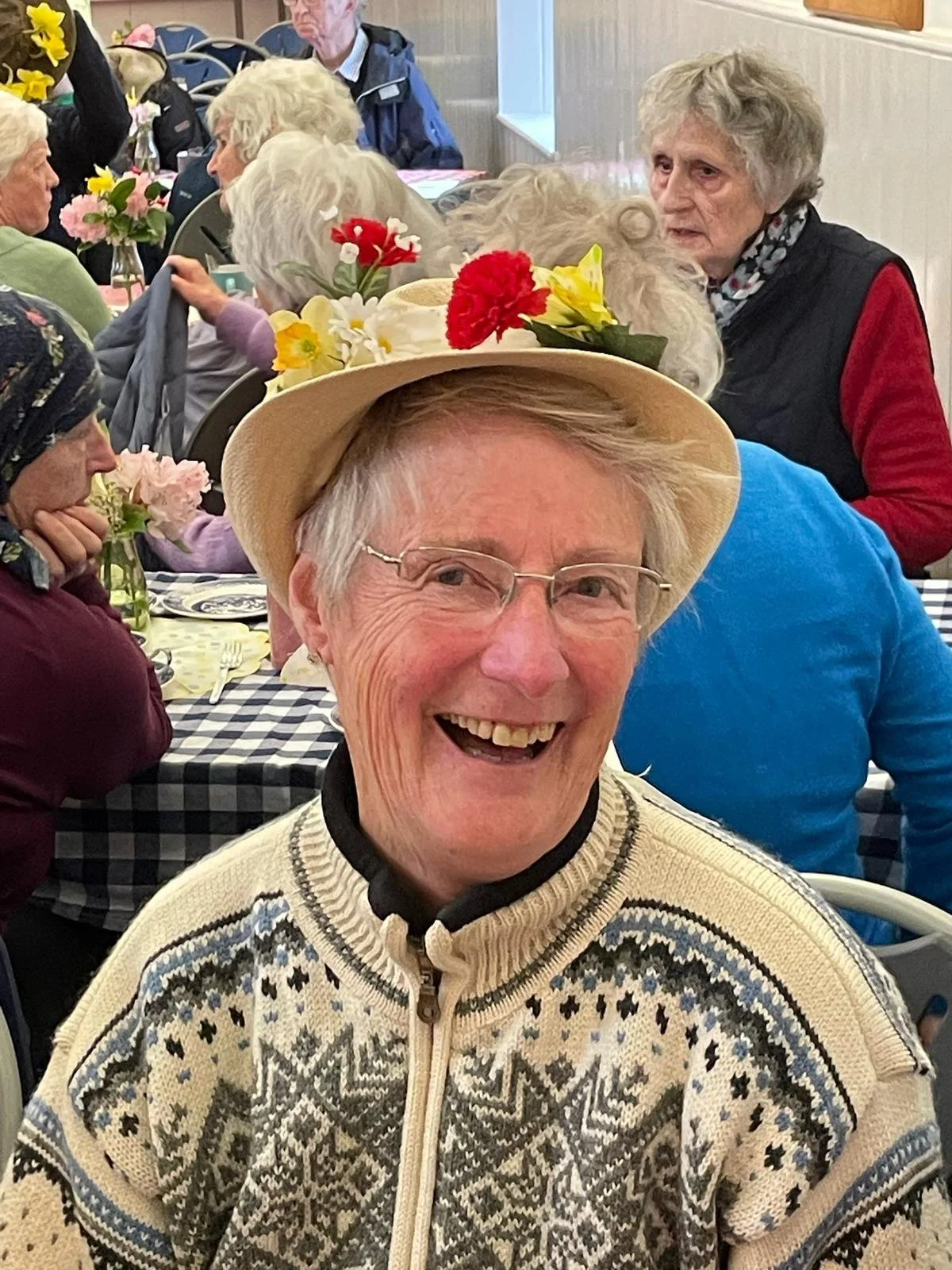 Smiling elderly woman wearing glasses and a beige hat decorated with colorful flowers, sitting at a table with a checkered tablecloth amidst a group of elderly people at a social gathering.