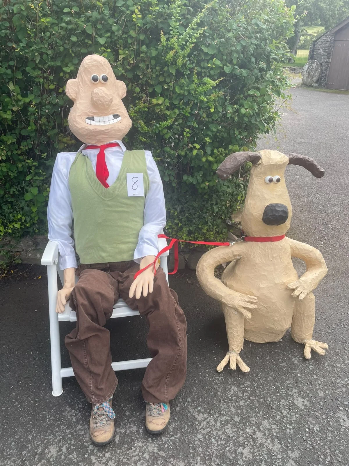 A large, smiling puppet figure with googly eyes, a big nose, wearing a light green vest, white shirt, red tie, and brown pants, is seated on a white outdoor chair. Beside it, a large paper-mâché dog puppet with floppy ears, googly eyes, a big black n