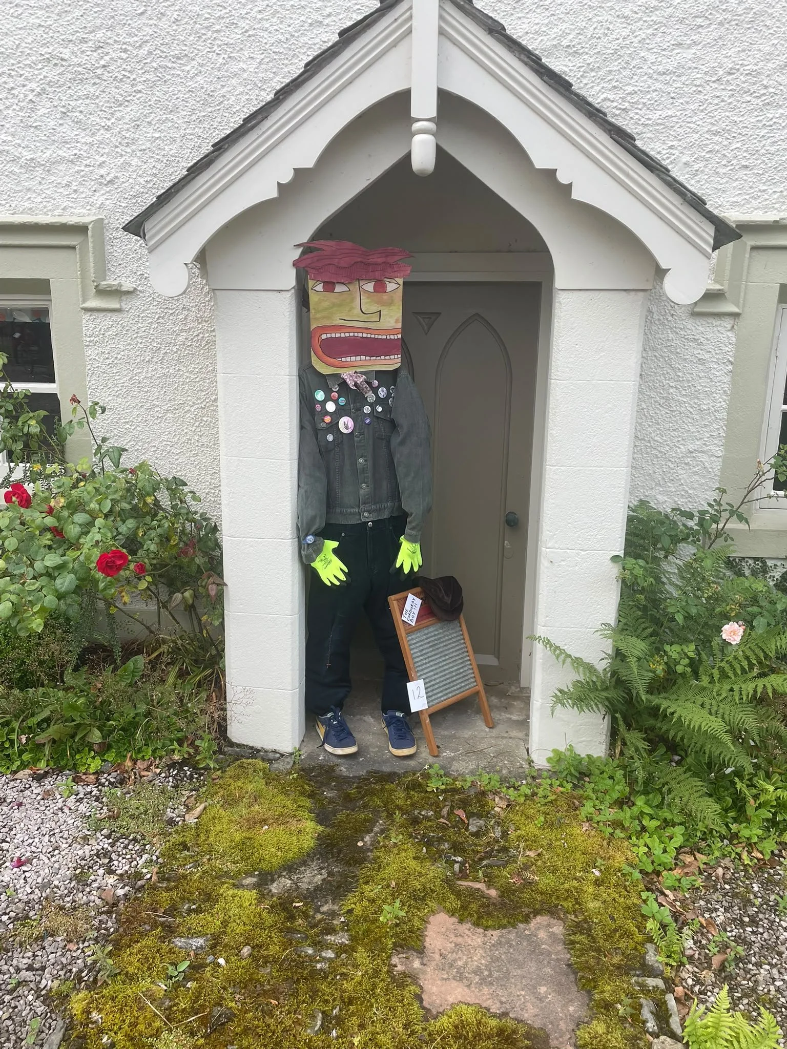 A person wearing a Halloween mask, yellow gloves, black pants, a gray jacket, and neon yellow shoelaces, standing in a small white porch with a triangular roof, surrounded by green plants and flowers. The porch has a wooden sign with the number 12 le
