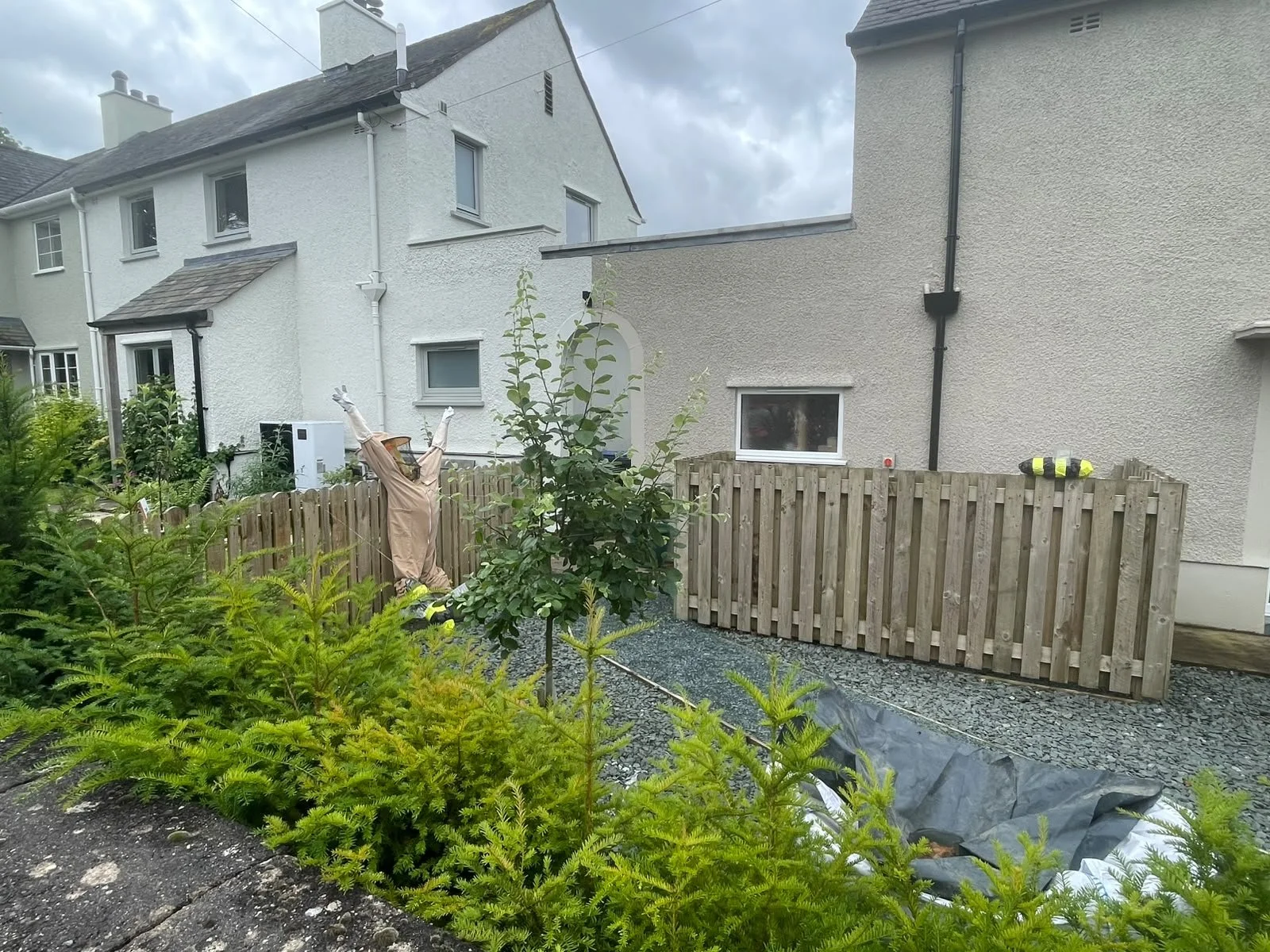 Residential backyard with a wooden fence, green plants, and a small tree. A scarecrow dressed in a beige suit and a wide-brimmed hat is placed near the fence. Two yellow safety vests are hanging on the fence. The background features white and beige h